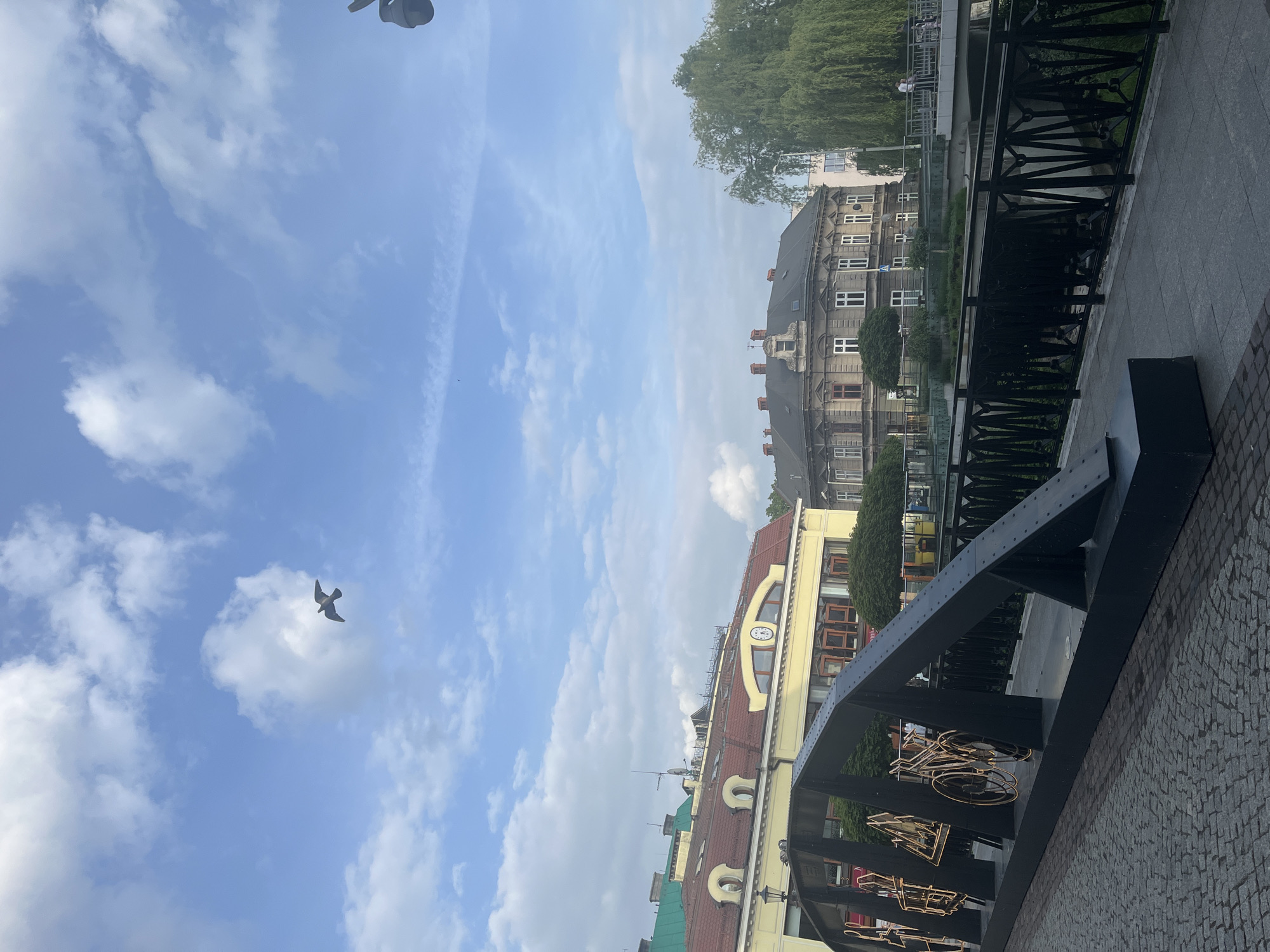 The image shows a cityscape under a bright sky with scattered clouds. In the foreground, a black metal bridge structure adorned with stylized bicycle shapes is visible. A stone-paved street is also visible. Adjacent to the bridge are buildings with varied architectural styles, including a yellow building with a clock on its facade and a taller, gray building with multiple windows. In the background, green trees and a canal or river can be seen. A bird is flying in the sky.
