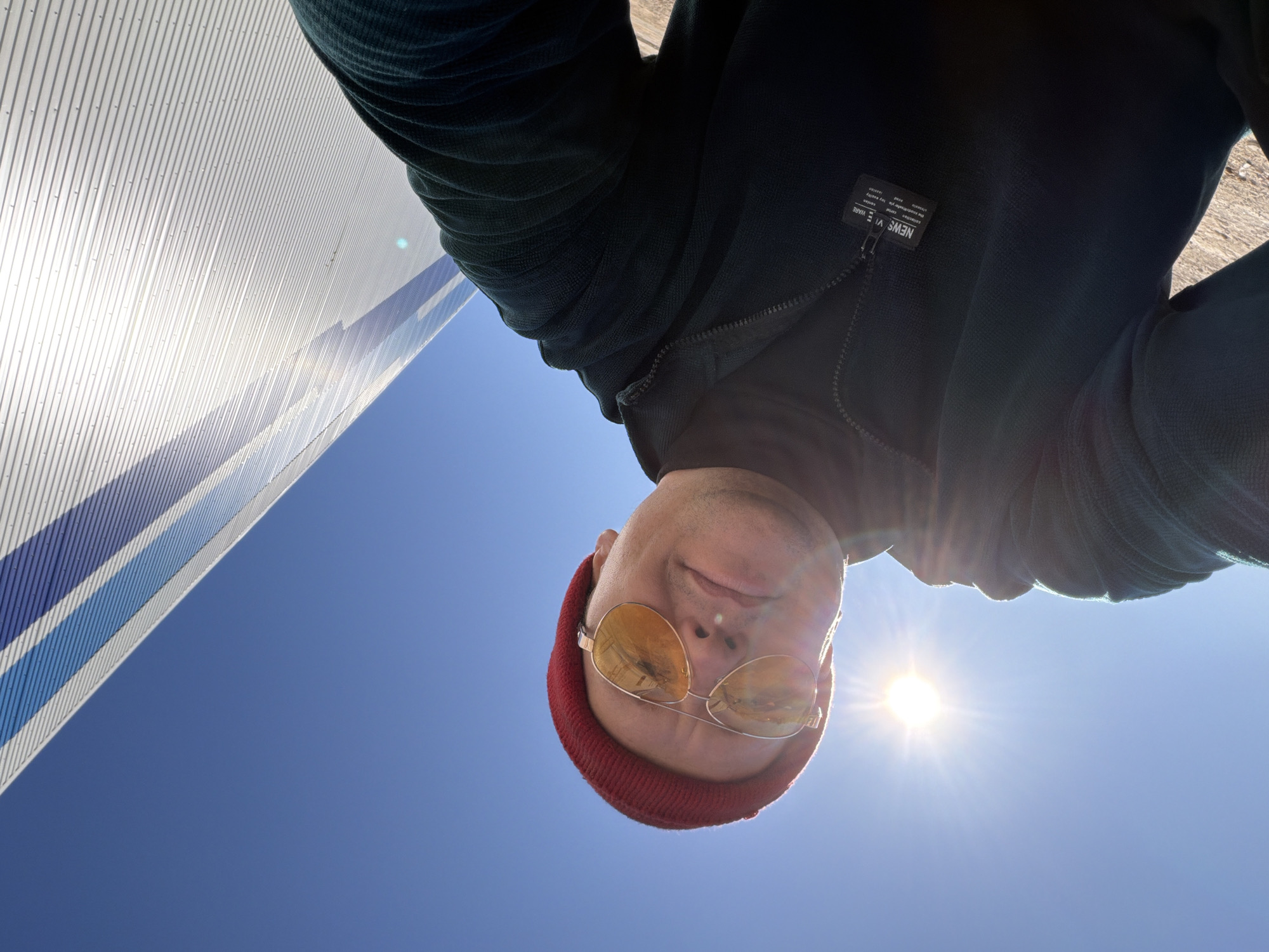A low-angle shot depicts a person wearing a red beanie and yellow-tinted sunglasses, positioned against a bright blue sky with the sun shining directly in the frame. The person is wearing a dark-colored zip-up jacket with a visible label on the left chest. A modern building with white and blue corrugated metal siding is partially visible in the upper left corner of the frame. The location is in the city of Chaikovsky, Russia.