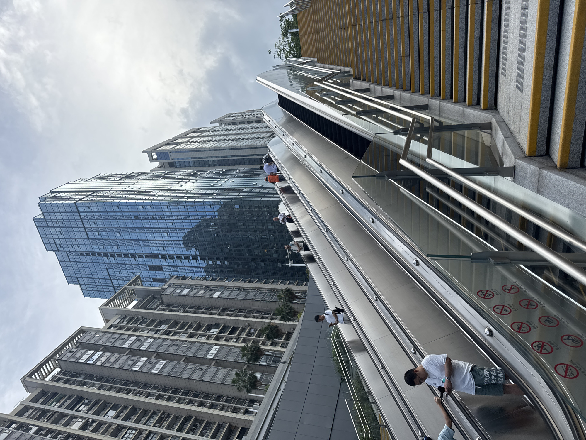 Captured at an extreme angle, the photograph showcases an outdoor escalator system in Futian District, China. The escalator is sandwiched between a series of staircases with yellow and grey accents and modern skyscrapers. Several individuals are riding the escalator in both directions, one notably wearing a white t-shirt and patterned shorts, holding onto the handrail. The skyscrapers have a glass and concrete facade and a light, overcast sky serves as the backdrop, emphasizing the urban landscape.