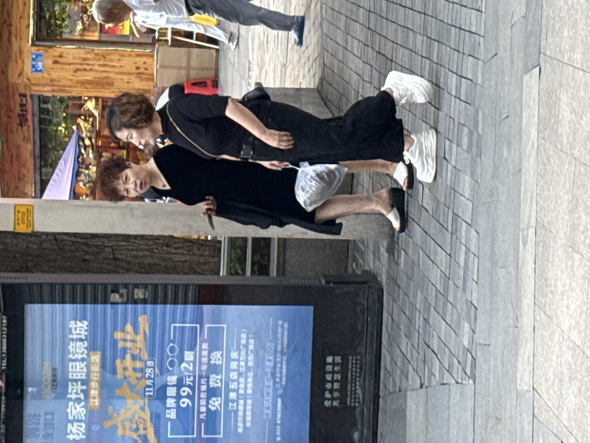 The image captures a street scene in Jiangjin District, Chongqing, China. Two adult women are the primary subjects. The woman on the left, with short, dark curly hair and a red hue, wears a black dress and white platform sandals. She holds a smartphone in her left hand and a plastic bag with items inside in her right. The woman on the right, with shoulder-length dark hair, is dressed in a black top with a black belt and black wide-leg pants. She wears white platform sneakers and has a dark crossbody bag. Both women are walking on a tiled sidewalk.
Behind the women, a large blue advertisement display is visible, featuring Chinese text and graphics related to an eyeglasses store named "Yangjiaping Eyeglasses City". The ad highlights a grand opening on November 28th, with promotions for branded eyewear at 99 yuan for two pairs and a free replacement service for children's anti-blue light lenses for one year. Further details on the ad mention a branch in Jiangjin Wudian, with addresses for various locations in the district.
The environment appears to be an urban setting with commercial establishments. To the right of the advertisement, a wooden-fronted building with signs is visible. A purple awning extends from a shopfront. Other individuals are present in the background, including a man in a white shirt and grey pants walking away from the camera and another person partially visible. The overall lighting suggests daytime.