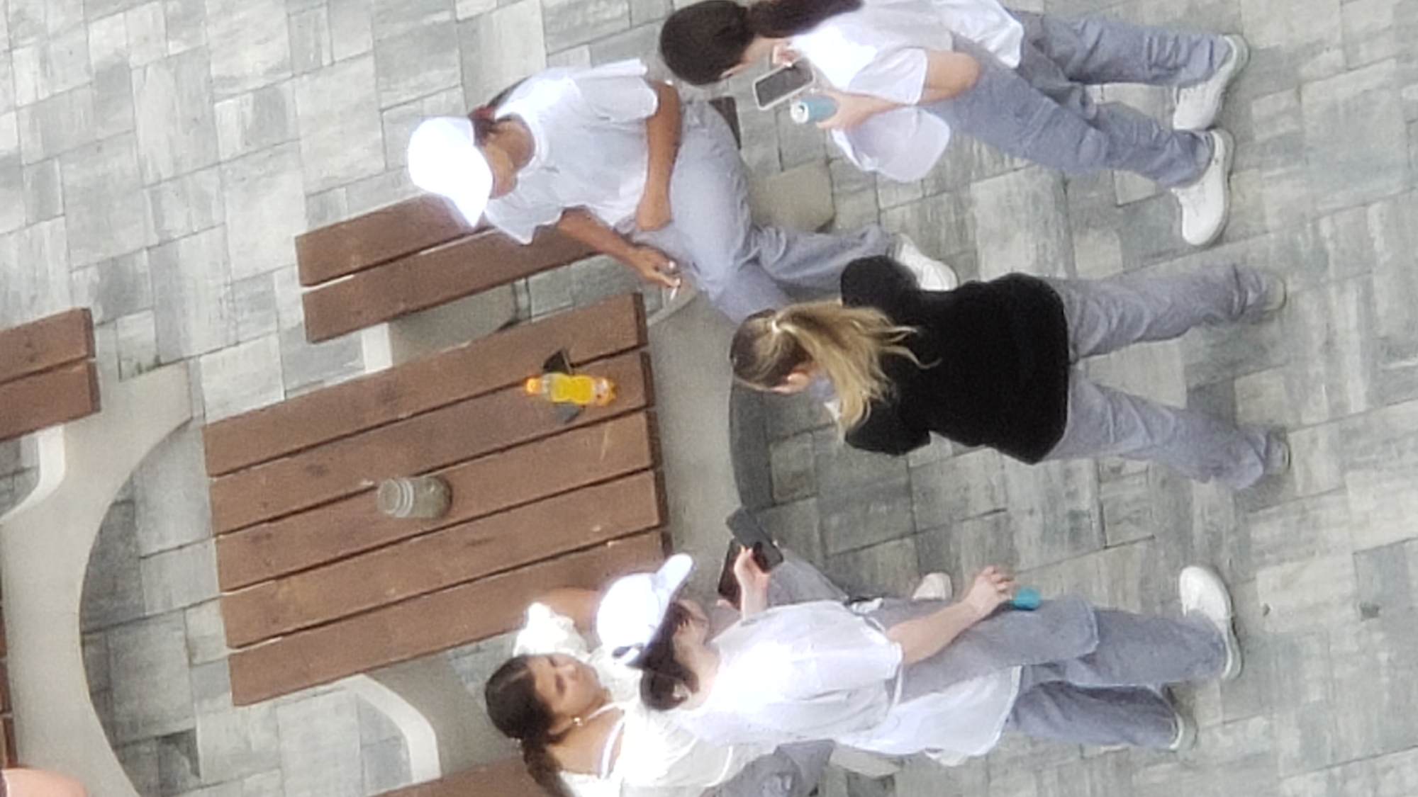 The image depicts a group of five individuals, appearing to be young women, gathered around a wooden picnic table outdoors. The setting is an urban environment with a paved patio area. The individuals are dressed casually in light-colored tops and pants, with some wearing white baseball caps.
One woman on the left is seated, wearing a white top and looking towards the center. To her right, another woman is standing, wearing a white top and a white cap, and is holding a phone. A bottle of orange liquid and a glass jar are visible on the table. A third woman is seated on the right side of the table, wearing a white t-shirt and light-colored pants, and appears to be smoking. She has a white cap on her head.
In the foreground, two women are standing. One is facing away from the camera, wearing a black top and light-colored pants, and holding a can. The other, also facing away, is wearing a white top and light-colored pants, and is holding a phone.
The environment suggests a public space, possibly a park or a recreational area within the city of Prešov, Slovakia, given the architectural style of the paving stones and the benches. The lighting indicates daytime.