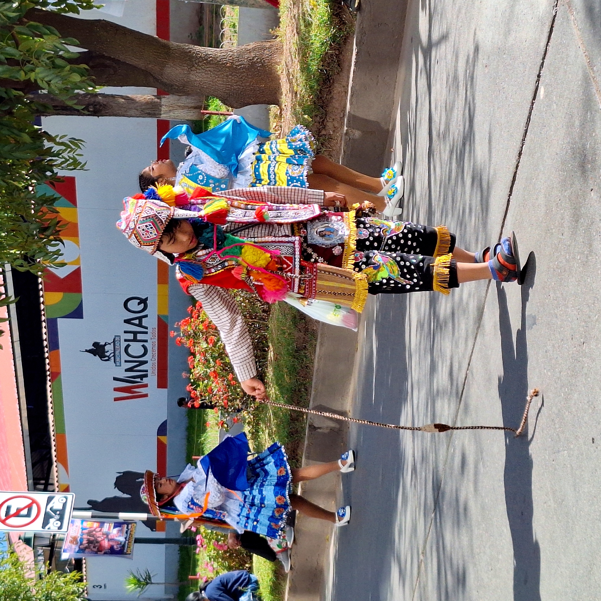 The image depicts a street scene in Wanchaq, Peru, likely during a parade or festival, as indicated by the traditional colorful attire of the individuals. In the foreground, a young person in a detailed costume, featuring a patterned hat with tassels, a vest adorned with embroidery and pom-poms, and patterned pants with fringed hems, walks forward, holding a braided whip. To their left, a girl in a white dress with blue accents and a blue skirt walks past, holding a blue fabric. To the far left, a child in a similar white and blue outfit is partially visible. In the background, a large white wall displays the text "MUNICIPALIDAD DE WANCHAQ Unidos Crecemos Todos" along with a silhouette of a horse and rider. A traffic sign with a red circle and diagonal line over an "E" symbol is also visible on the left. Red flowers border the street, and trees with green foliage provide shade. The ground is paved with asphalt, and shadows from the trees are cast upon it.