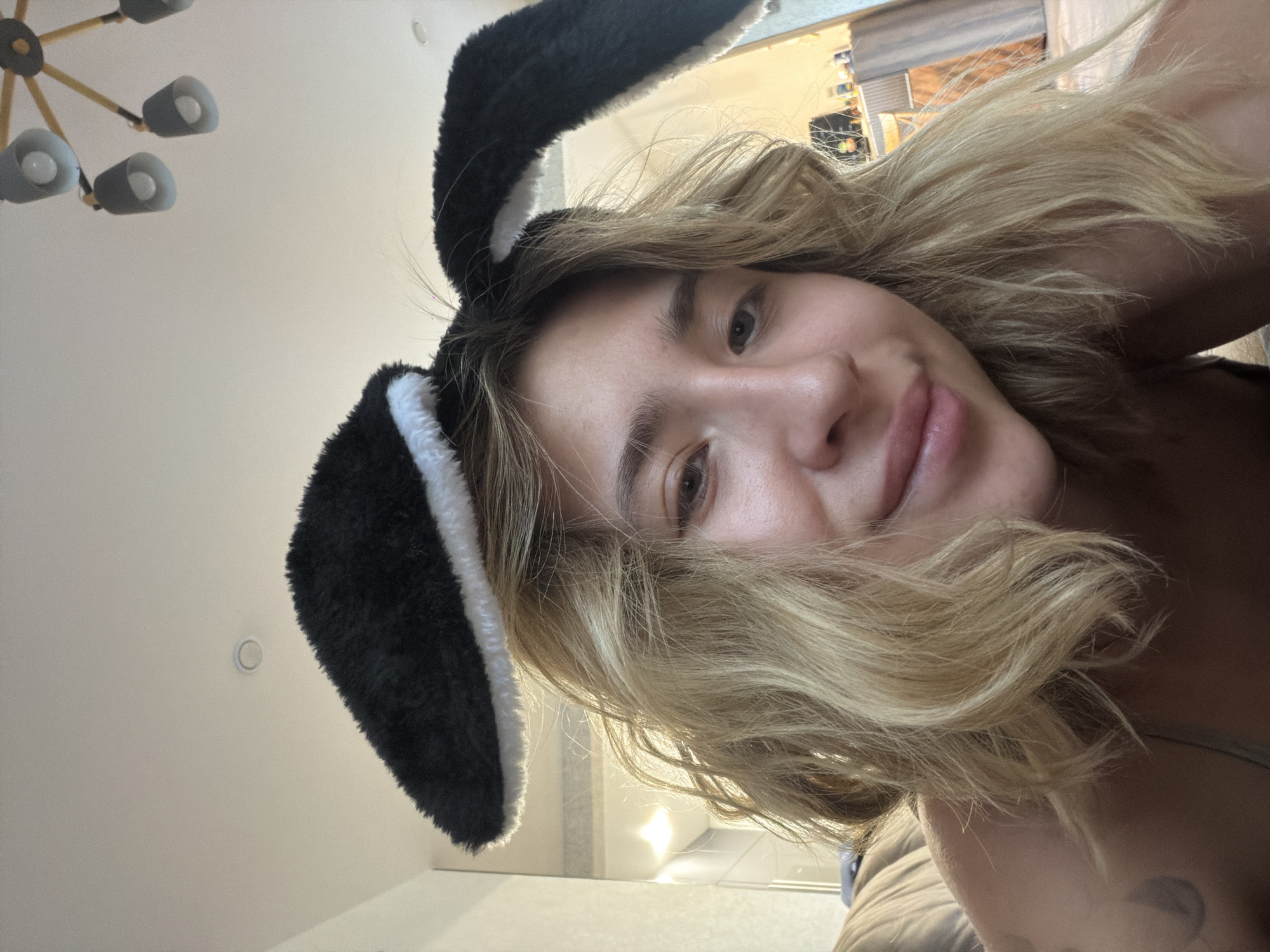 A young woman with blonde, wavy hair and light skin is shown from the chest up, taking a selfie. She is wearing black and white faux fur bunny ears on her head. The background appears to be an indoor setting, possibly a bedroom, with a neutral-colored wall and a glimpse of a chandelier on the ceiling. There are also reflections in a mirror or glass surface on the left side of the image. The overall lighting suggests it is daytime, with natural light entering the room. The exact location within Kamenka, Russia, cannot be determined from the visual information provided.