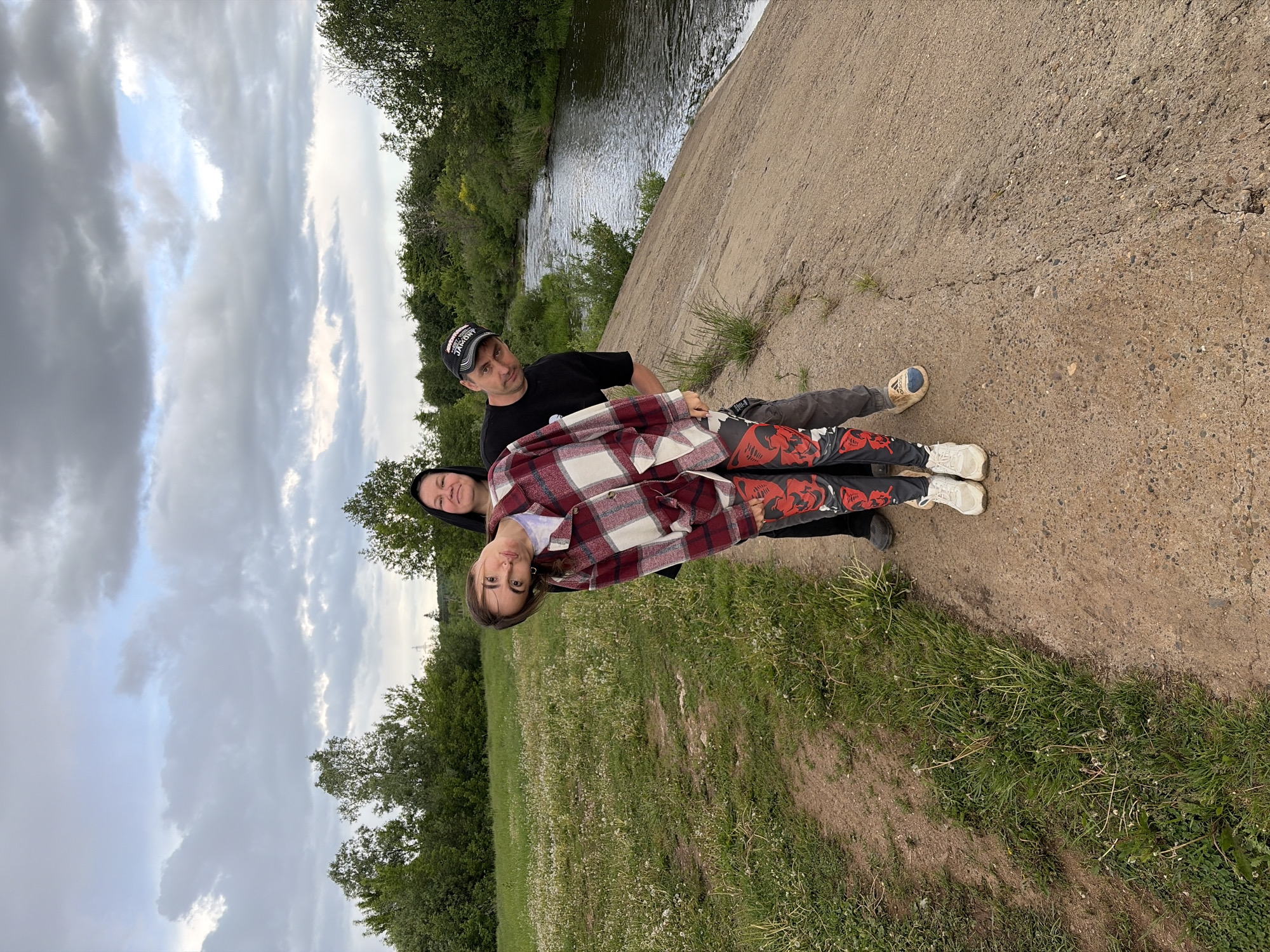 The image shows three people outdoors near a body of water, possibly a river. A man in a black t-shirt and a baseball cap is standing on a concrete structure, slightly behind a woman wearing a black hooded garment. In front of them is a younger woman wearing a red and white plaid jacket and pants with a graphic design. They appear to be posing for a photo. The location is identified as Nizhnii Vishur, Russia. The background includes trees, grass, and a cloudy sky.