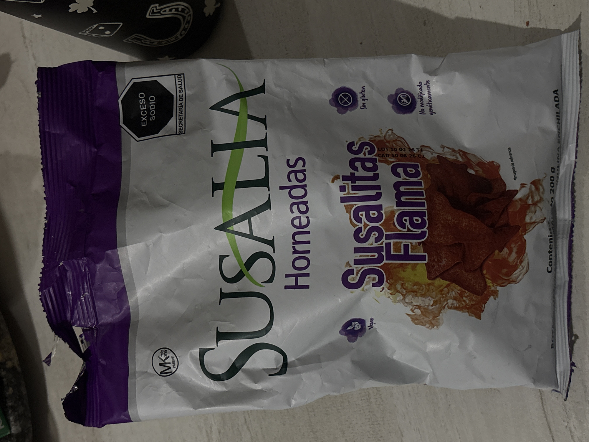 A partially opened 200g bag of "SUSALIA Hornoadas Susalitas Flama" snack chips is positioned centrally on a light-colored, textured surface. The bag is white with a purple top and displays a black octagonal "EXCESO SODIO" (Excess Sodium) warning from the "SECRETARÍA DE SALUD" (Ministry of Health) on the upper right. Other visible markings include "MK PAREVE" (Kosher Pareve), "Vegano" (Vegan), "Sin gluten" (Gluten-free), and "No modificado genéticamente" (Not genetically modified) labels. The bag features an illustration of red/orange chips with a fiery effect. To the right of the bag, a portion of a black object adorned with white designs such as stars, shamrocks, and horseshoes is visible. The scene is located in San Bernardino Tlaxcalancingo, Mexico.