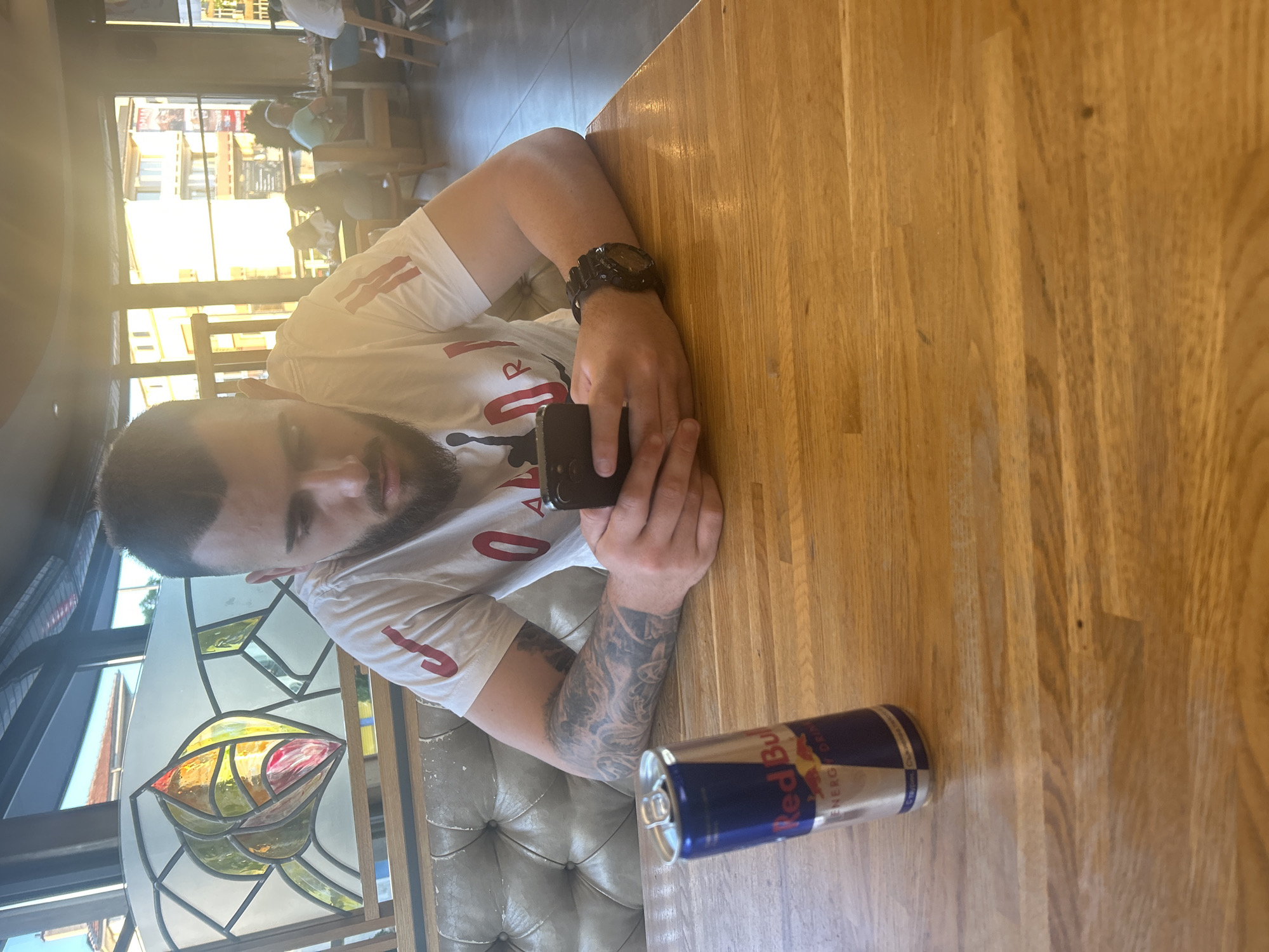 A man with dark hair and a beard is seated at a wooden table in what appears to be a cafe or restaurant. He is wearing a white t-shirt with red lettering and a large black watch on his left wrist. He is holding a black smartphone in his hands and looking at it. An open can of Red Bull energy drink is on the table near his arm. Behind him is a stained glass window. Other patrons and tables are visible in the background outside the window. The location is Kazanlak, Bulgaria.