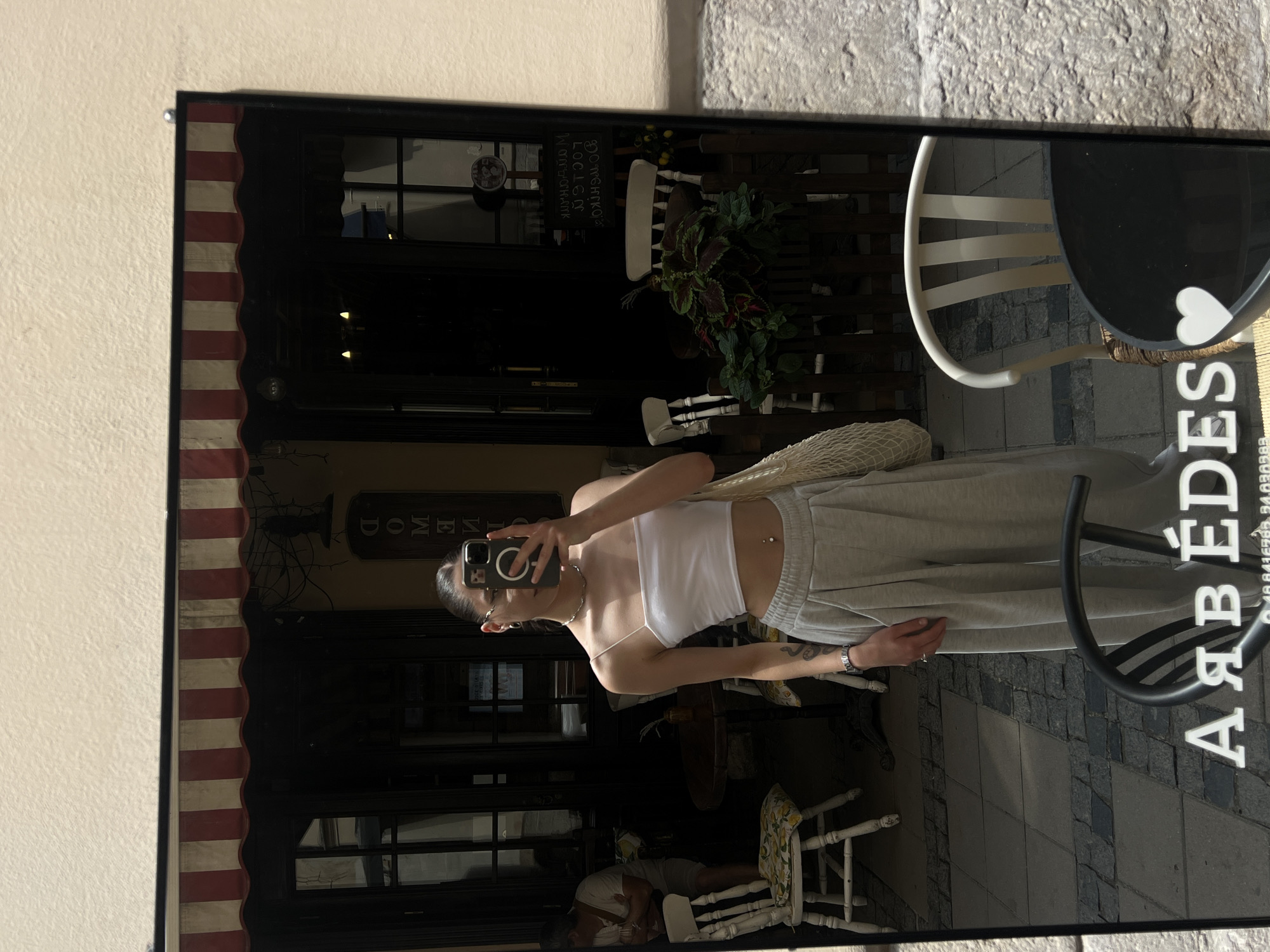 The image is a mirror selfie taken by a young woman in Lviv, Ukraine. She is wearing a white crop top and light grey sweatpants. She has her hair tied up in a bun and is holding a smartphone to take the picture.
In the reflection, we can see the woman standing on a cobbled street, likely in front of a cafe or restaurant. The cafe has a red and white striped awning and large glass doors. There are tables and chairs visible both inside and outside the cafe. Some of the chairs are white, while others appear to be made of wood. There are also potted plants and a decorative fence. On the ground, there is a sign with white lettering that says "AARBEDES".
The lighting suggests it is daytime. The overall scene depicts a casual street-side setting in a European city.