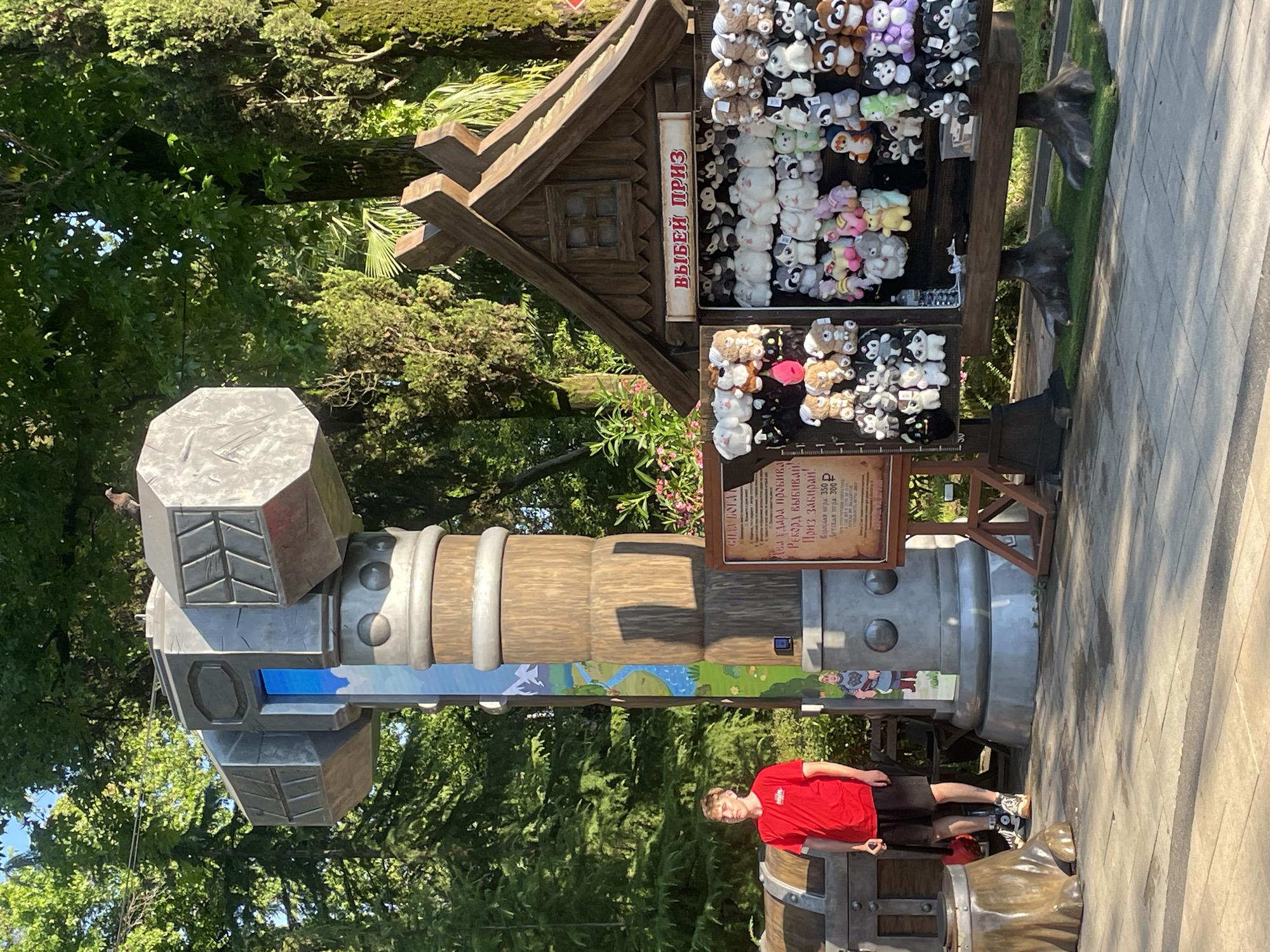 The image depicts an outdoor scene in Sochi, Russia, likely at an amusement park or entertainment venue. In the foreground, a young man in a red t-shirt and black shorts stands to the left, holding a dark-colored pole. To his right is a large, stylized hammer prop, resembling Mjolnir from Norse mythology, with a blue panel in the center. Behind the hammer, a wooden stall displays a variety of plush toys, arranged on shelves. The stall has a sign with Russian text that reads "ВЫБЕЙ ПРИЗ," meaning "Pick a Prize." The background features lush green trees and foliage, indicating a warm climate. A small bird is perched on top of the hammer prop. A treasure chest prop is visible to the left of the young man. The ground is paved with light-colored rectangular stones.