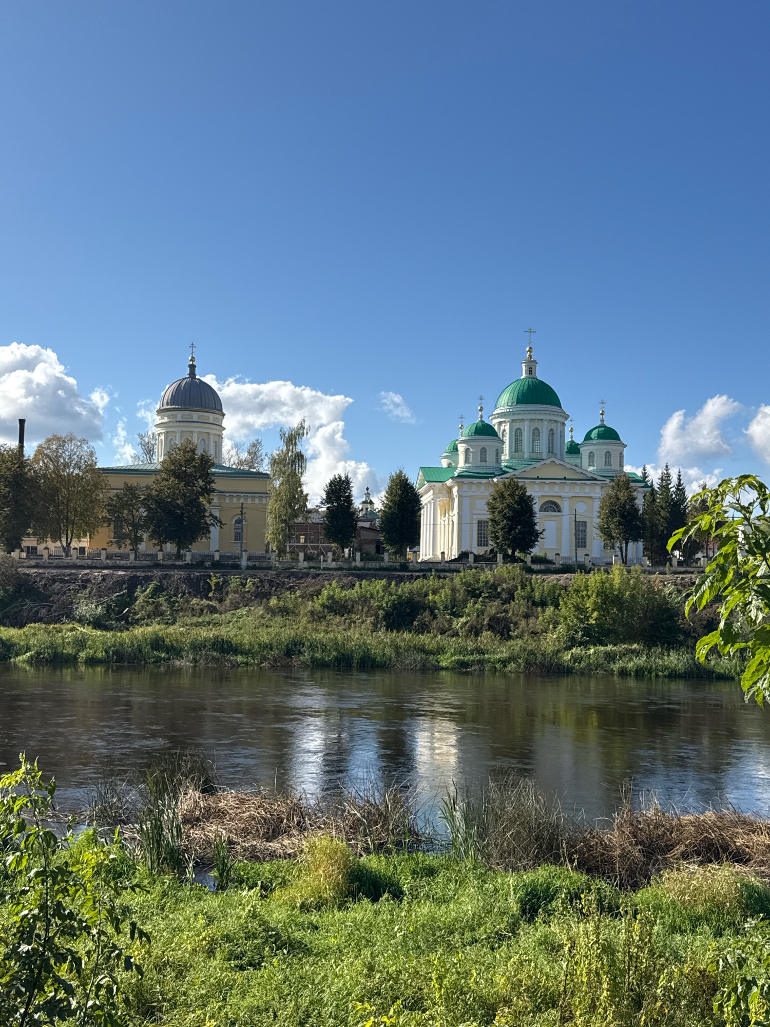 The image depicts a serene landscape featuring two prominent religious structures situated on the bank of a wide, calm river. The larger, more elaborate building on the right is a cathedral with multiple green domes, white columns, and ornate architectural details. To its left, a slightly smaller, older-looking structure with a single, dark dome and a yellow facade stands. The river in the foreground reflects the sky and the buildings. The riverbank is lush with green vegetation, including tall grasses and trees. The sky is a vibrant blue, dotted with fluffy white clouds. The scene suggests a peaceful, sunny day in a historically rich location. This scene is likely the Borisoglebsky Monastery in the city of Torzhok, Russia, renowned for its picturesque setting and historical significance.