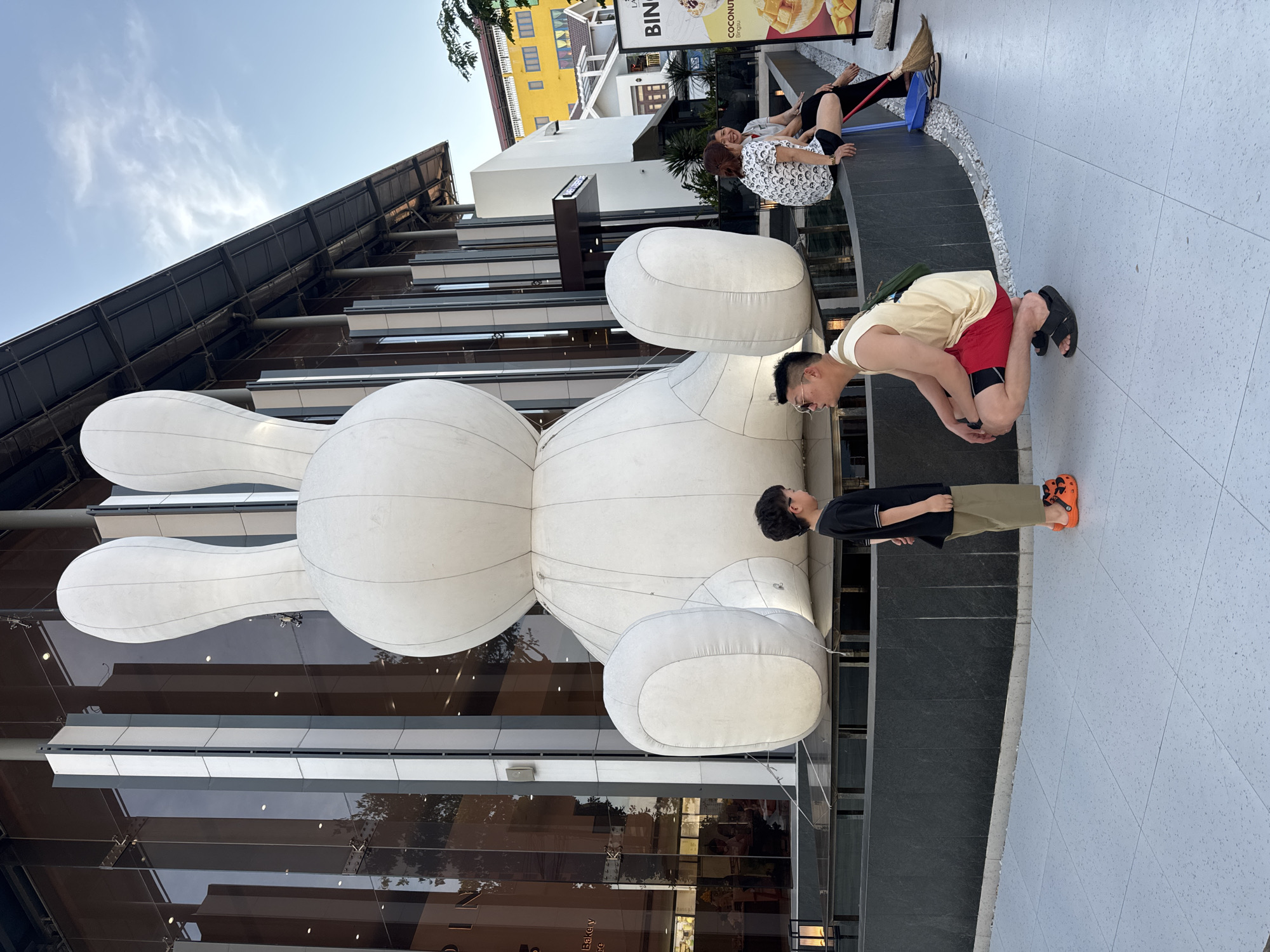 Two men stand beside a large white inflatable rabbit sculpture outside a modern building with reflective glass facades. One man, wearing a yellow tank top and red shorts, bends over with his hands near his knees. The other man, wearing a black shirt and khaki pants, stands beside him. Two women sit on a ledge with a broom and other cleaning items. Behind them is a building with a sign that says "BIN". The location is Đà Nẵng, Vietnam. The weather appears bright and sunny.