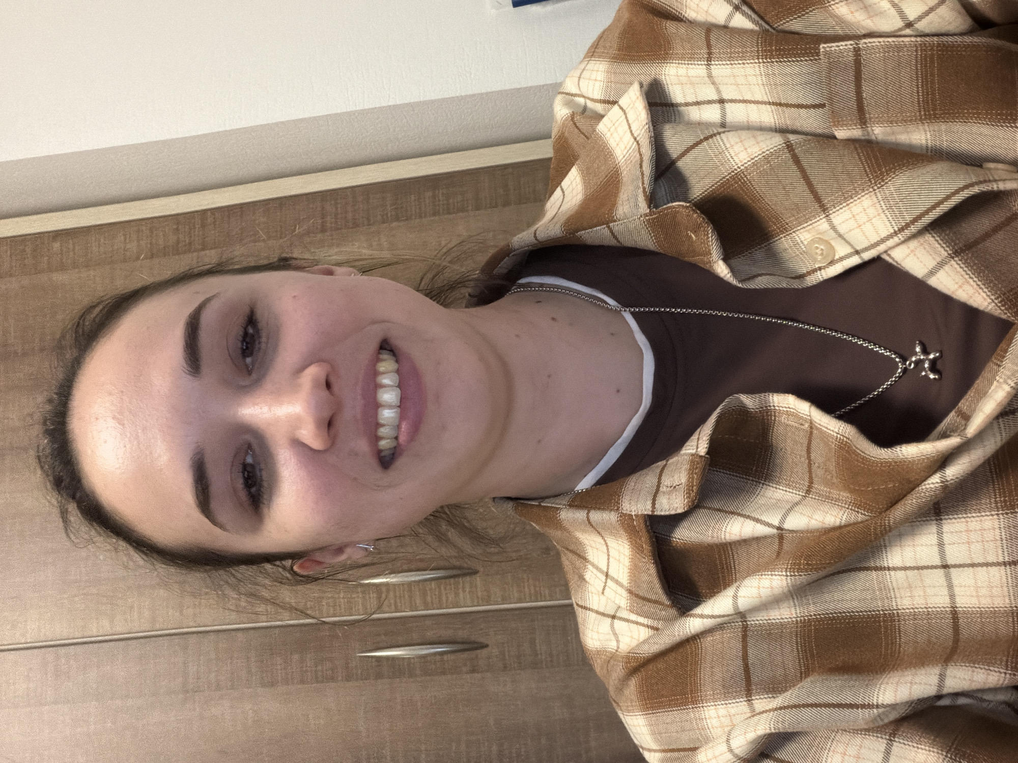 A young woman is facing the camera, smiling. She is wearing a brown t-shirt with a white neckline and a brown and white plaid flannel shirt, which is unbuttoned and draped loosely around her. A silver chain necklace with a pendant is visible on her chest.
The background consists of a light-colored wall and what appears to be a wooden cabinet or drawer.
The image does not provide enough information to establish the location as Rostov-on-Don, Russia.