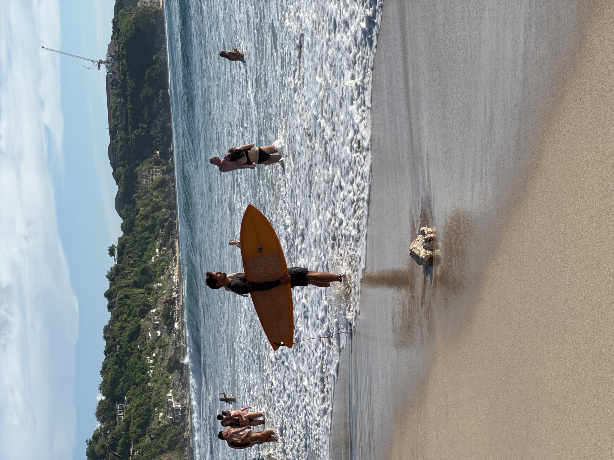 This image captures a vibrant beach scene in Kuta Selatan, Indonesia, on a clear, sunny day. The view is oriented horizontally, showcasing a sandy shore, the ocean, and a verdant cliffside.
In the foreground, the beige sand transitions from dry to wet as it meets the foamy white surf of the ocean. A small, light-colored rock or piece of coral rests on the wet sand on the left. The breaking waves create a thick layer of white foam that extends across much of the mid-ground.
The primary subject is a man standing in the shallow, foamy water on the left, holding an orange surfboard against his body. He wears a dark shirt and shorts and has a beard. His back is mostly turned towards the camera, and he appears to be looking out towards the ocean, possibly preparing to surf or having just finished. A red leash extends from his surfboard into the water.
Further into the water, several other individuals are visible. On the left, a group of people, including women in bikinis, are standing or wading in the foamy water. Another person in dark swimwear is seen further out in the more open water, seemingly swimming or floating.
The background is dominated by a lush, green, tree-covered cliff or hillside that runs along the coastline. Scattered among the dense foliage are several white or light-colored buildings, possibly hotels or residences, indicating human development. On the far left, a tall red and white construction crane is prominent against the blue sky, suggesting ongoing development in the area. The sky above the cliff is a bright blue with a few scattered white clouds.
The scene depicts a leisurely beach environment, with people enjoying the ocean. No visible text is present in the image.