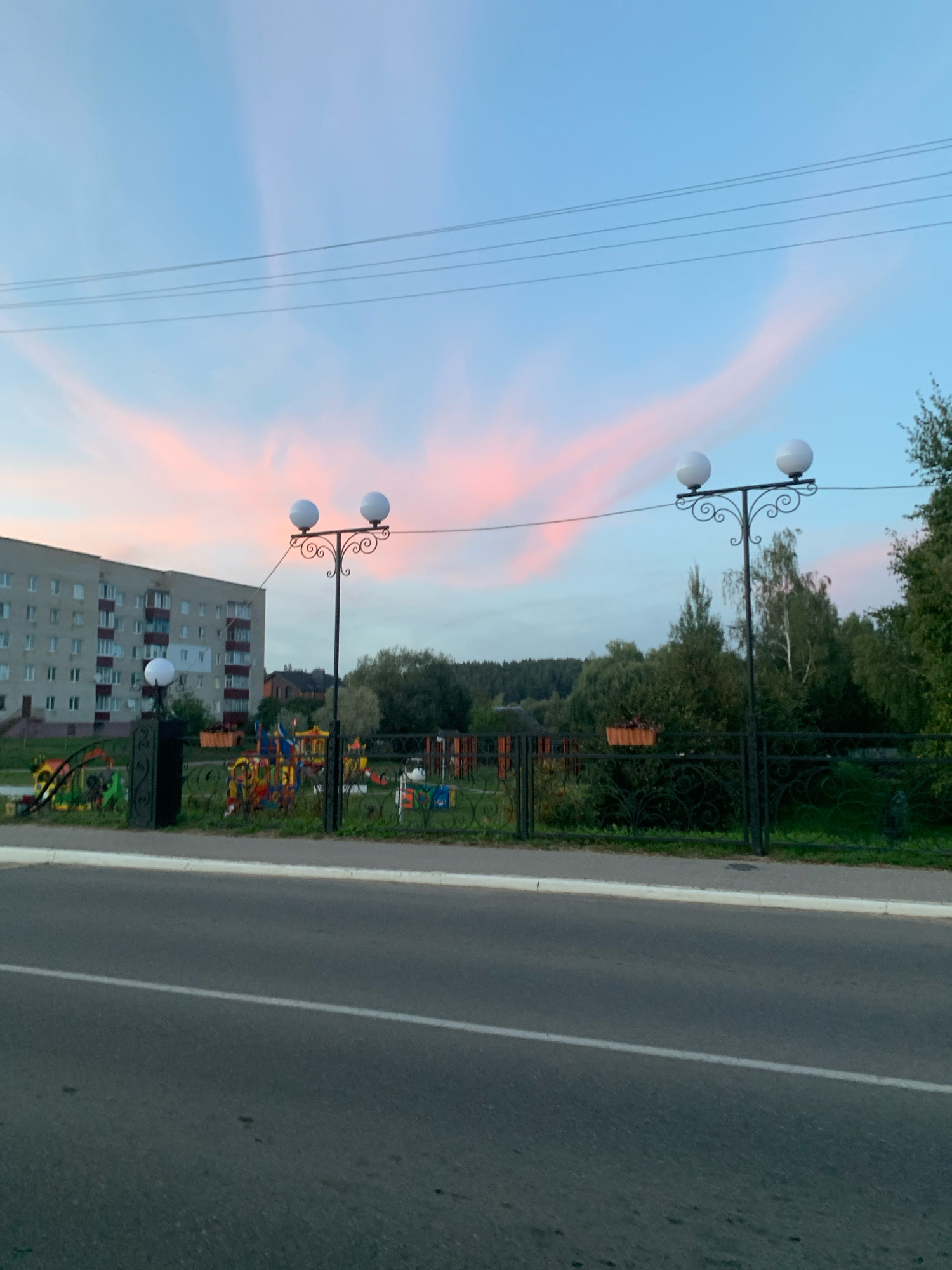 The image shows a street view in Kapyĺ, Belarus, at dusk. In the foreground, an asphalt road with a white dividing line stretches across the bottom of the frame. Beyond the road is a sidewalk and a black wrought-iron fence. Through the fence, a children's playground with colorful equipment is visible, situated in front of a multi-story apartment building. The sky above is a pale blue transitioning to pink and orange hues, indicating the setting sun, with thin, wispy clouds. Two ornate lampposts with spherical white lights stand along the fence line. Trees and vegetation are present in the background, forming a natural border to the residential area. Overhead power lines are also visible against the sky.