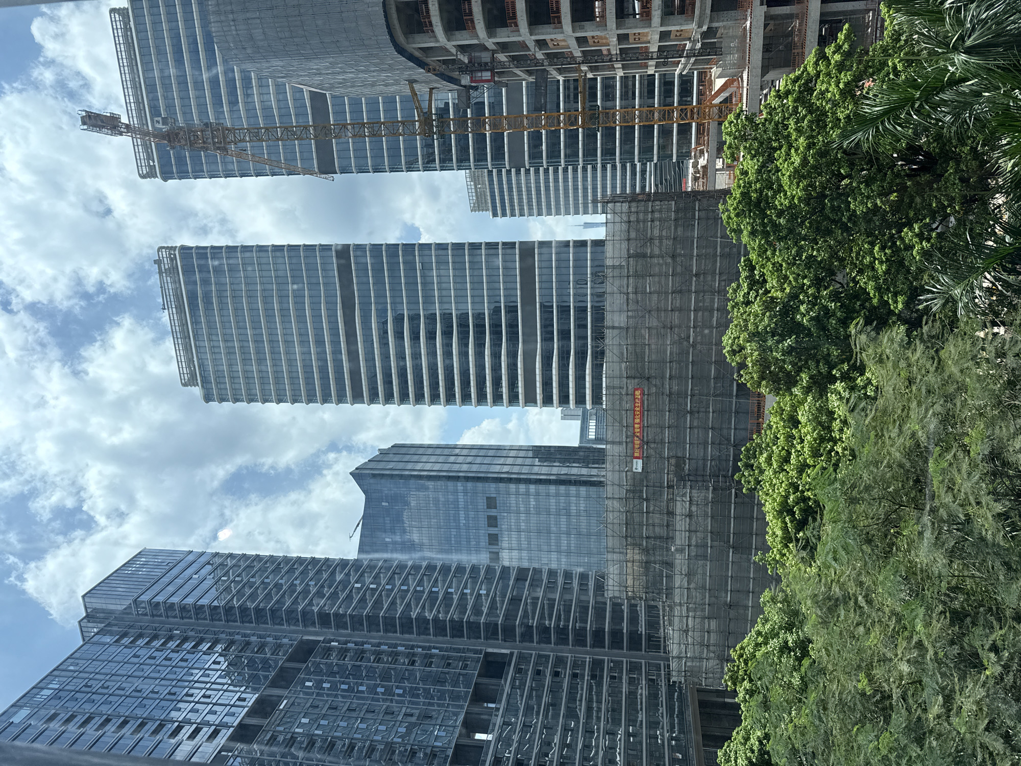 The image displays a cityscape in Haizhu District, China, dominated by modern high-rise buildings and ongoing construction. In the foreground, lush green trees and foliage create a natural contrast to the urban development. Several towering skyscrapers, characterized by their glass facades, are visible, some completed and others appearing to be under construction.
A prominent feature of the scene is a construction site in the mid-ground, covered by scaffolding and netting, suggesting significant building activity. A tall yellow construction crane stands to the right of the frame, actively engaged in the building process. One of the buildings under construction shows exposed concrete structures and visible scaffolding. In the distance, the sky is partly cloudy, with white clouds against a blue background. A red banner with Chinese text is visible on the construction site.