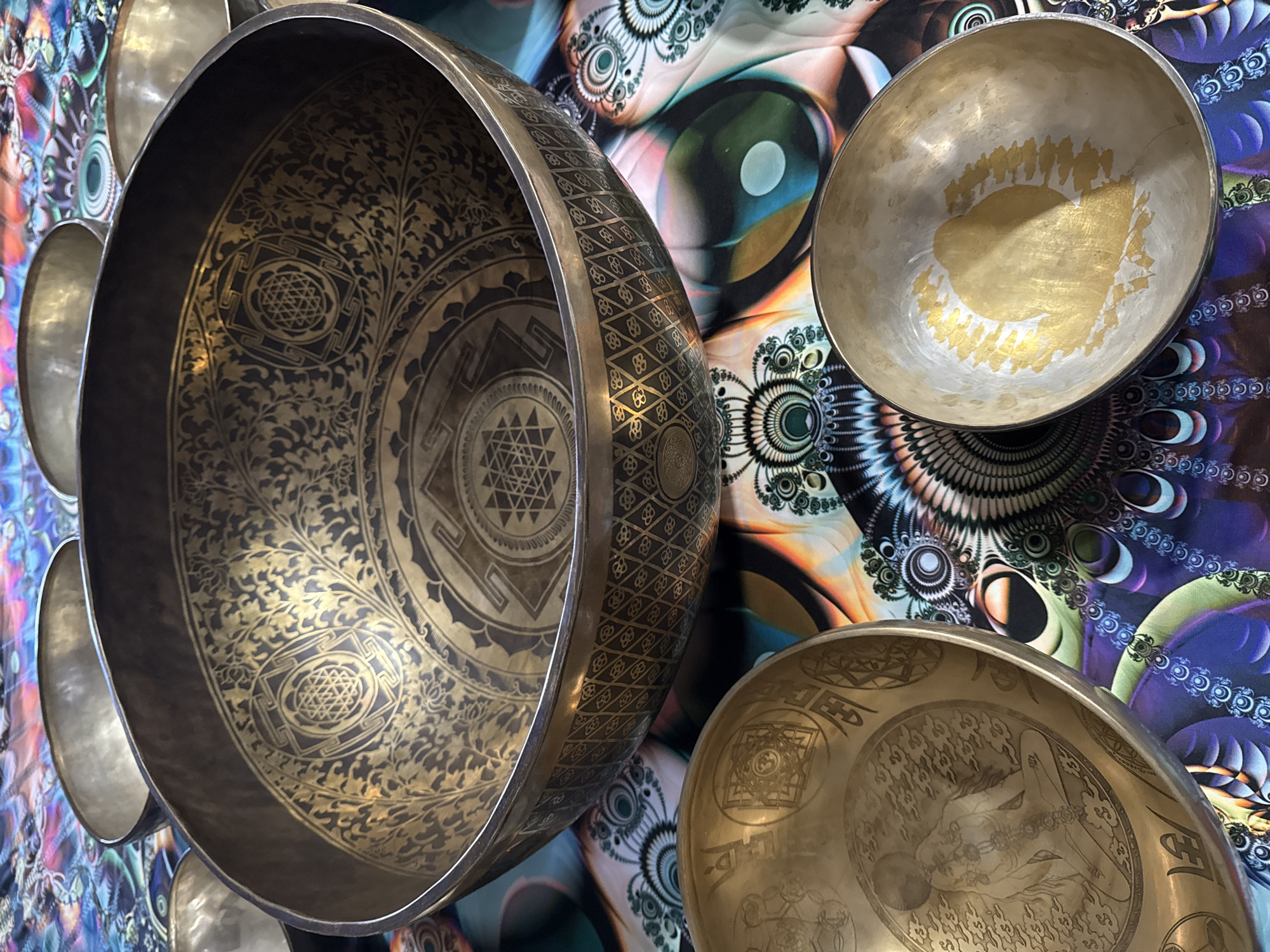 The image shows a collection of metal singing bowls resting on a patterned fabric. The bowls are various sizes and colors, ranging from silver to golden-bronze. Some of the bowls feature intricate designs etched into their surfaces, including geometric patterns and depictions of figures. The background fabric has a psychedelic, swirling pattern with vibrant colors. The location of this image is not included within this image. The image is most likely taken inside of a shop or private home.