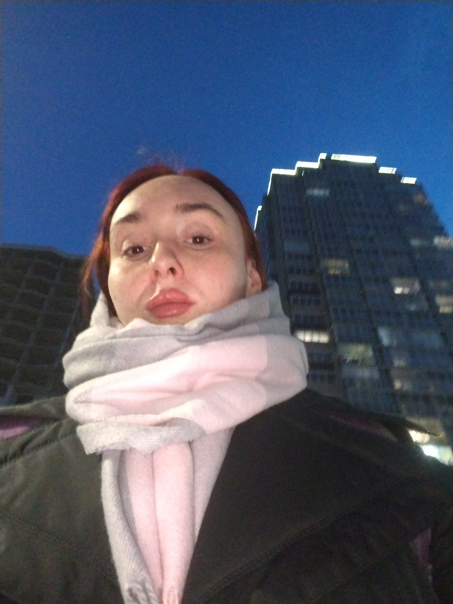 A low-angle selfie shows a woman with fair skin, red hair, and full lips, wearing a black jacket and a light gray and pink scarf. Behind her, a tall, dark building with lit windows rises into a dark blue sky. Another building is partially visible on the left. Given the architectural style and details, the location appears to be Izhevsk, Russia.