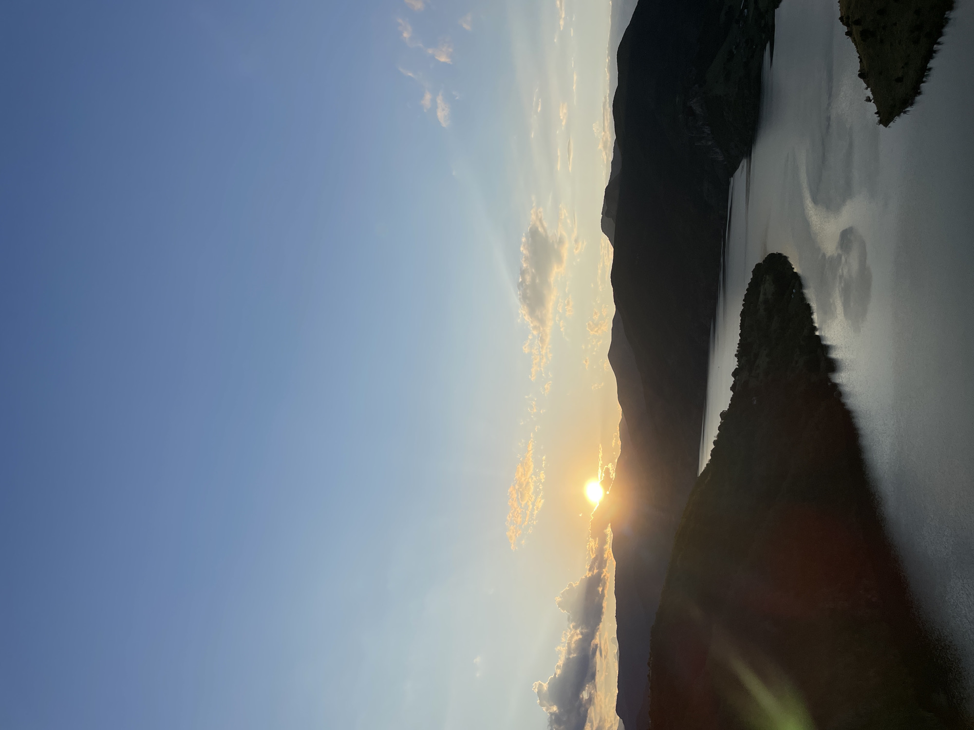 This is a vertical, eye-level shot of a sunset over a lake and mountains in the vicinity of Borovitsa, Bulgaria. The sun is setting behind the dark, silhouetted mountains, casting a warm glow on the clouds and the sky. The lake reflects the sky and mountains, creating a serene and peaceful atmosphere. The mountains in the foreground are covered in dark vegetation, while the mountains in the background are more rugged and mountainous. The sky is a clear blue with a few wisps of clouds.