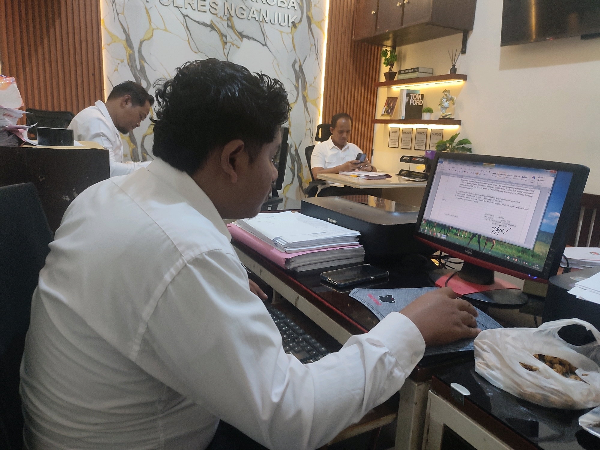 The image depicts an indoor office environment, likely within the city of Ringinanom, Indonesia, given the visible text "POLRES NGAN..." which refers to a police department. In the foreground, a man wearing a white long-sleeved shirt is seated at a desk, facing right, with his left arm extended towards a keyboard. His attention is directed towards the computer monitor in front of him, which displays a document. To his right, on the desk, is a white plastic bag containing snacks and a grey fabric item with a black design. Behind him, another man in a white shirt is seated at a desk, looking at his smartphone. Further back, a third man, also in a white shirt, is seated at a desk, appearing to work. The office walls have a decorative marble-like pattern and vertical wooden paneling. Shelving units are visible in the background, displaying books and decorative items. The overall scene suggests a professional workspace with individuals engaged in office tasks.