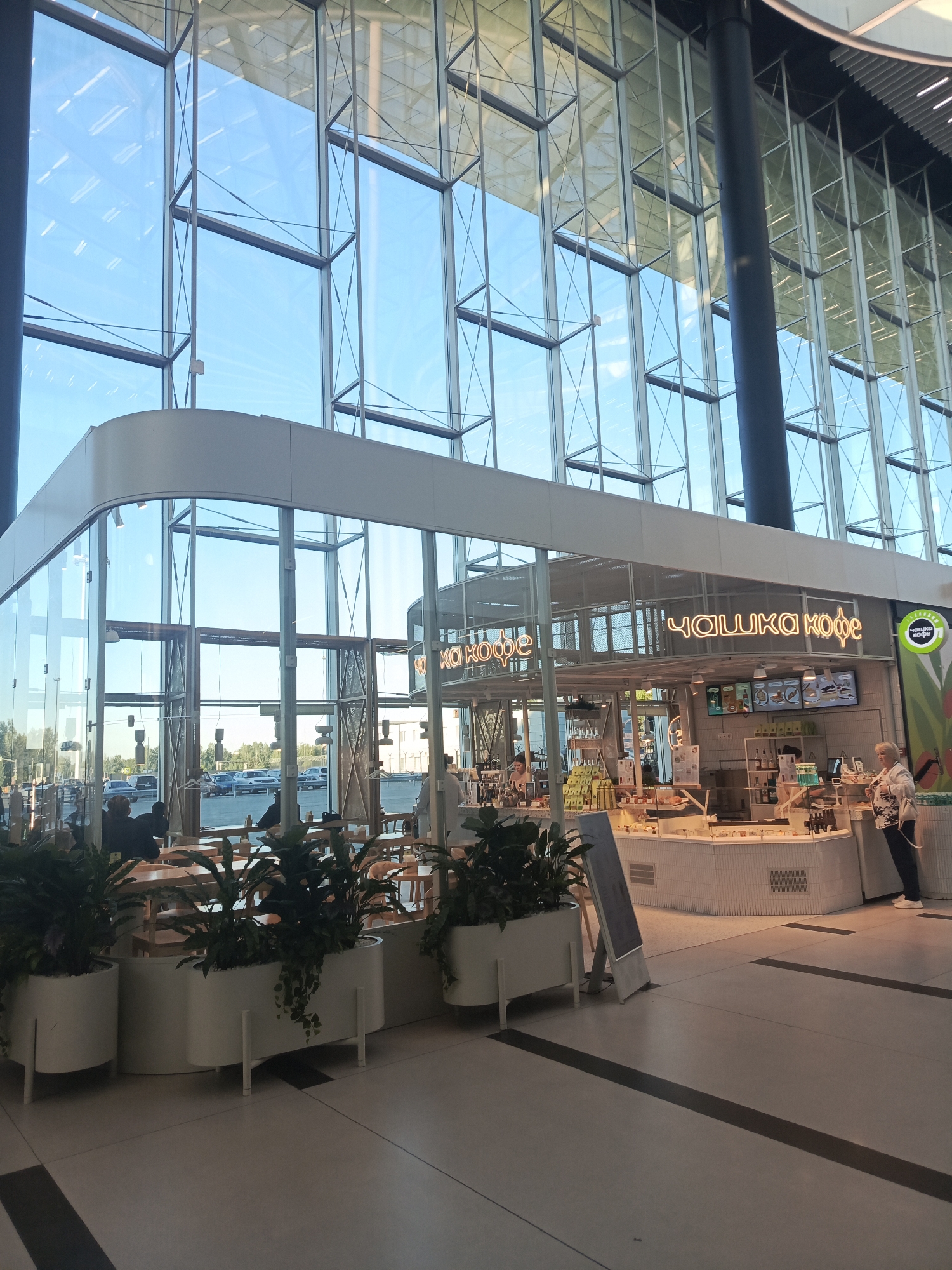 The image captures a contemporary cafe called "Чашка Кофе" (Chashka Kofe) situated within a modern building featuring a large glass facade. The cafe has a clean, white aesthetic with neon signs displaying the cafe's name. Inside, staff members are visible behind a counter stocked with food items and beverages. Customers are seated at tables in the foreground, and some are standing near the counter. Large planters filled with greenery are placed in front of the cafe, adding a touch of nature to the indoor space. Through the glass walls, a street scene with cars and trees is visible, suggesting an urban setting. The architectural style of the building, with its expansive glass and metal framework, points to a commercial or public building like a shopping mall or airport. The location is identified as Ob, Russia.