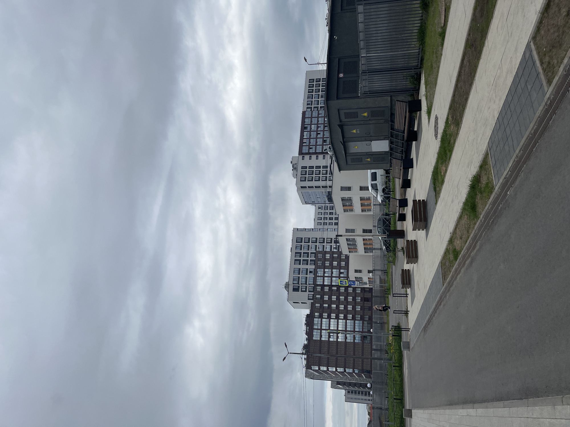 The image depicts a street in Saint Petersburg, Russia, lined with modern apartment buildings. The sky is overcast. A paved road runs alongside a pedestrian walkway, punctuated by benches and greenery. A person is walking along the sidewalk, next to a parking sign. Electrical infrastructure is visible adjacent to the buildings.