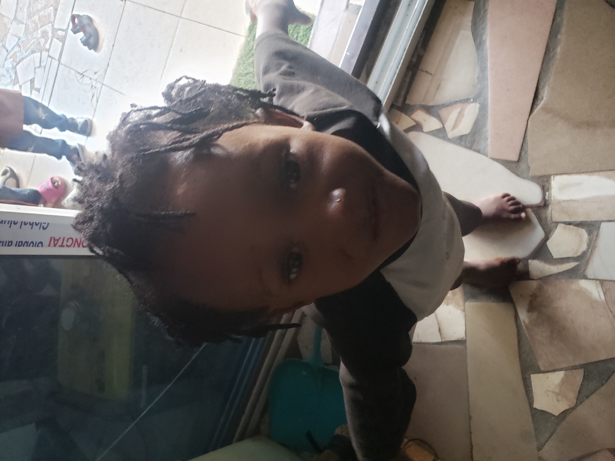 The image, taken in Adankolo, Nigeria, features a young child with dark skin and braided hair (possibly locs or cornrows) looking directly at the camera. The child, barefoot and wearing a white t-shirt over a dark long-sleeved garment, is positioned in the foreground, viewed from a slightly elevated perspective. Their feet rest on a floor composed of irregularly shaped, light-colored tiles, interspersed with darker patches.
The setting appears to be an indoor or semi-sheltered area, likely a shop entrance or a similar transitional space, brightly lit by natural daylight. A strip of artificial green turf is visible near the child's right foot. In the background, on the left, the lower legs of another person are visible, wearing dark trousers and dark shoes or sandals. A pair of pink slippers also lies nearby. A single red and black sandal is on the tiled floor further back.
To the left of the child, a glass surface, possibly a display case or window, features inverted text. Portions of the text visible include "ONGTAI Global alu...", suggesting a commercial entity, possibly related to "CHONGTAI Global Aluminum" or similar. A blue plastic dustpan or container is partially visible beneath the child. The child's expression is neutral and observant.