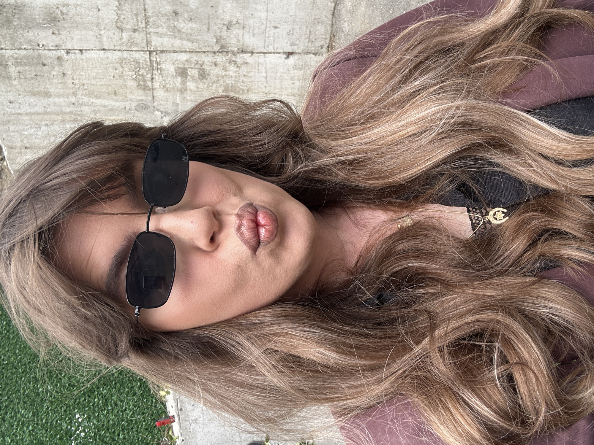 This close-up, angled portrait features a person, likely female, posing outdoors in Tbilisi, Georgia. The subject's head is tilted to the right, and her lips are playfully pursed in a "kiss" expression. She wears dark, rectangular sunglasses that partially obscure her face. Her long, wavy hair, a mix of light brown and blonde highlights, flows down her shoulders and chest. She appears to be wearing a dark top, possibly with lace details, underneath a brown or maroon jacket or overshirt. A delicate gold necklace with a star and crescent moon pendant is visible on her chest. The background elements include a textured concrete surface on the left and upper portion of the image, and a patch of green artificial turf or grass on the lower left. The lighting indicates it is daytime. No text is visible in the image.
