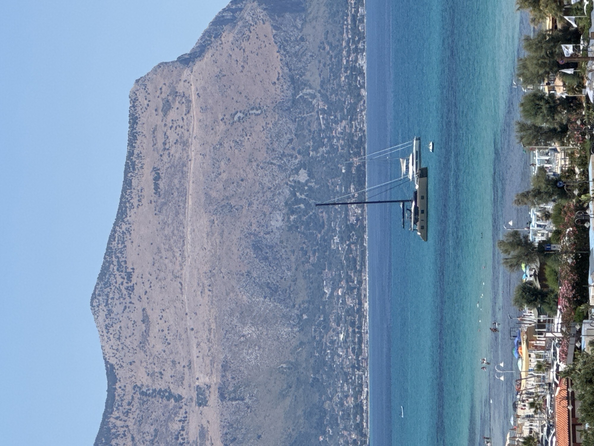 This media file captures a bright, sunny day at Isola delle Femmine, Italy. The scene is dominated by a towering, rugged mountain on the left, its slopes covered in sparse, dry vegetation and dotted with darker patches of shrubbery. To the right of the mountain, a picturesque coastline unfolds. The sea is a vibrant turquoise, transitioning to a deeper blue further out, and it appears calm.
Several boats are visible on the water. In the mid-ground, a large sailboat with white sails and a beige hull is moored, and a smaller white boat is nearby. Closer to the shore, numerous small white objects, possibly buoys or other small boats, are scattered. The shoreline itself is lined with buildings, suggesting a small town or village. Trees and some flowering bushes add greenery and color to the waterfront.
In the water near the shore, several people can be seen enjoying the day. Some are swimming or wading, while others appear to be engaged in water activities. A few small boats are also closer to the shore, and there are people on them. The overall atmosphere is one of leisure and a beautiful summer day at the coast. The clear blue sky indicates fair weather. There is no visible text in the image.
