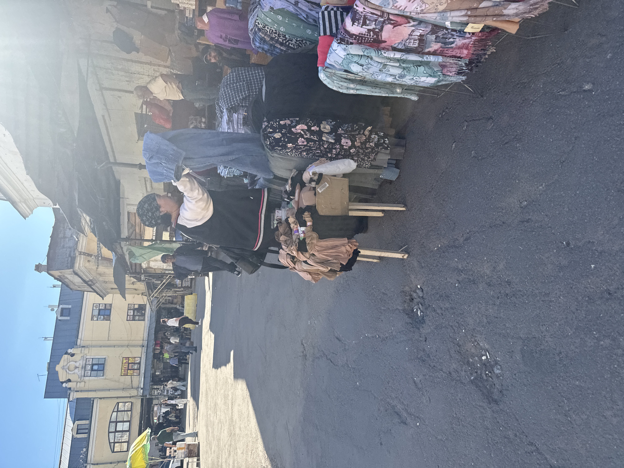 A street market is depicted in Saint Petersburg, Russia. In the foreground, a person wearing a black jacket with white and red stripes, a white shirt, and a patterned headscarf is holding up a pair of light blue jeans. Behind them, a stall displays various folded clothes and textiles, including patterned scarves and blouses in shades of purple, blue, green, and pink. Another stall to the right is filled with more clothing items.
Further in the background, several other people can be seen browsing or working at the market. The architecture of the buildings suggests an urban environment. The buildings are light yellow with dark blue roofs and arched windows. Signs with Cyrillic text are visible on the buildings, one of which reads "ПУЛЬС 35". The sun is shining, casting shadows on the asphalt ground, indicating it is daytime. The overall atmosphere seems to be that of a busy marketplace on a clear day.
