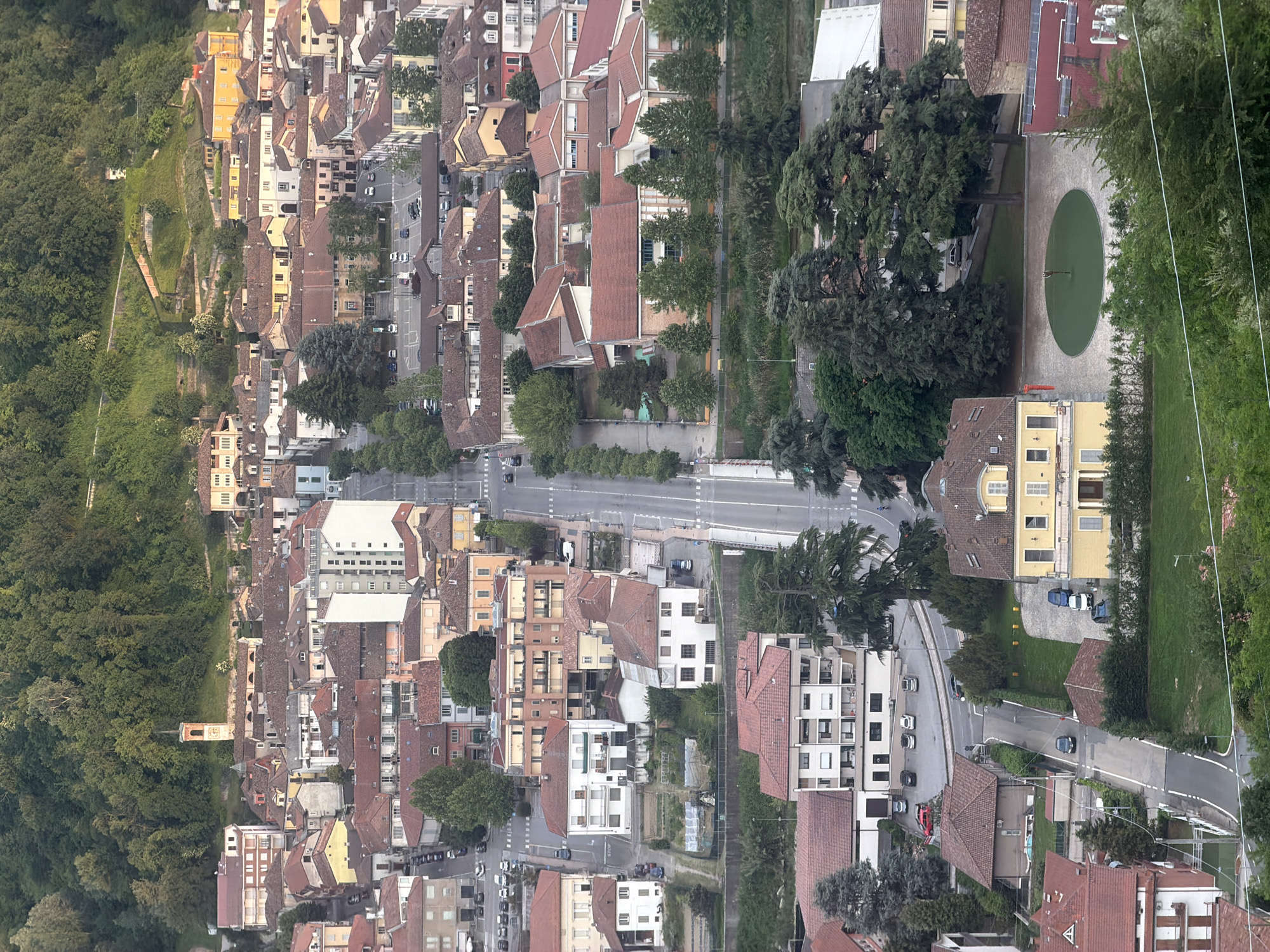 The image shows an aerial view of a section of Santo Stefano Belbo, Italy. At the upper left is a densely wooded hillside. Adjacent to the woods is a row of closely packed buildings with red tile roofs. A main road runs horizontally through the center of the image, with buildings and trees lining both sides. To the right, a circular area with a green surface is surrounded by trees and buildings. Cars are seen parked and driving on the roads. The overall environment is a mix of residential and commercial buildings interspersed with green spaces.