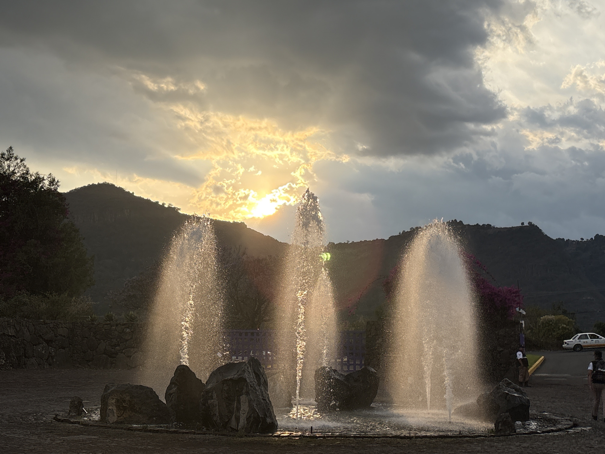 The image captures a fountain with three prominent jets of water shooting upwards. Large, dark rocks surround the base of the fountain. The backdrop consists of a mountain range silhouetted against a sky partially obscured by dramatic, grey clouds, with the setting sun peaking through, casting a golden light. In the distance, a white car is visible to the right, and two people are standing near the edge of the fountain. The scene appears to be in San Sebastian, Mexico.