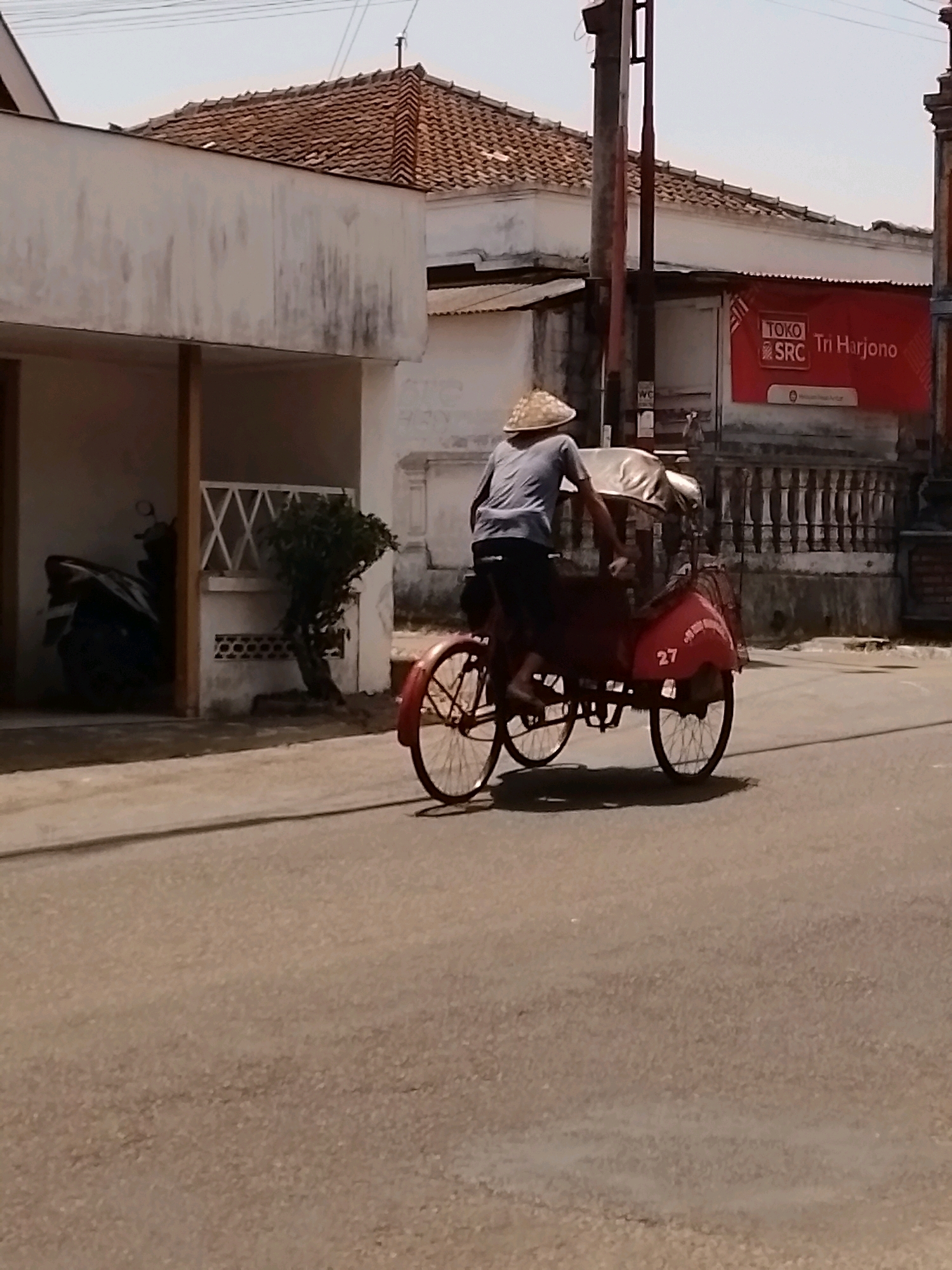A man wearing a conical hat is riding a red, three-wheeled pedicab on a paved road in front of buildings in Wonosari, Indonesia. The pedicab has a covered passenger area and the number "27" is visible on its rear. Behind the pedicab is a white building with a red sign that reads "TOKO SRC Tri Harjono". Another building with a tiled roof is visible to the left. The sky is clear and the lighting suggests it is daytime.