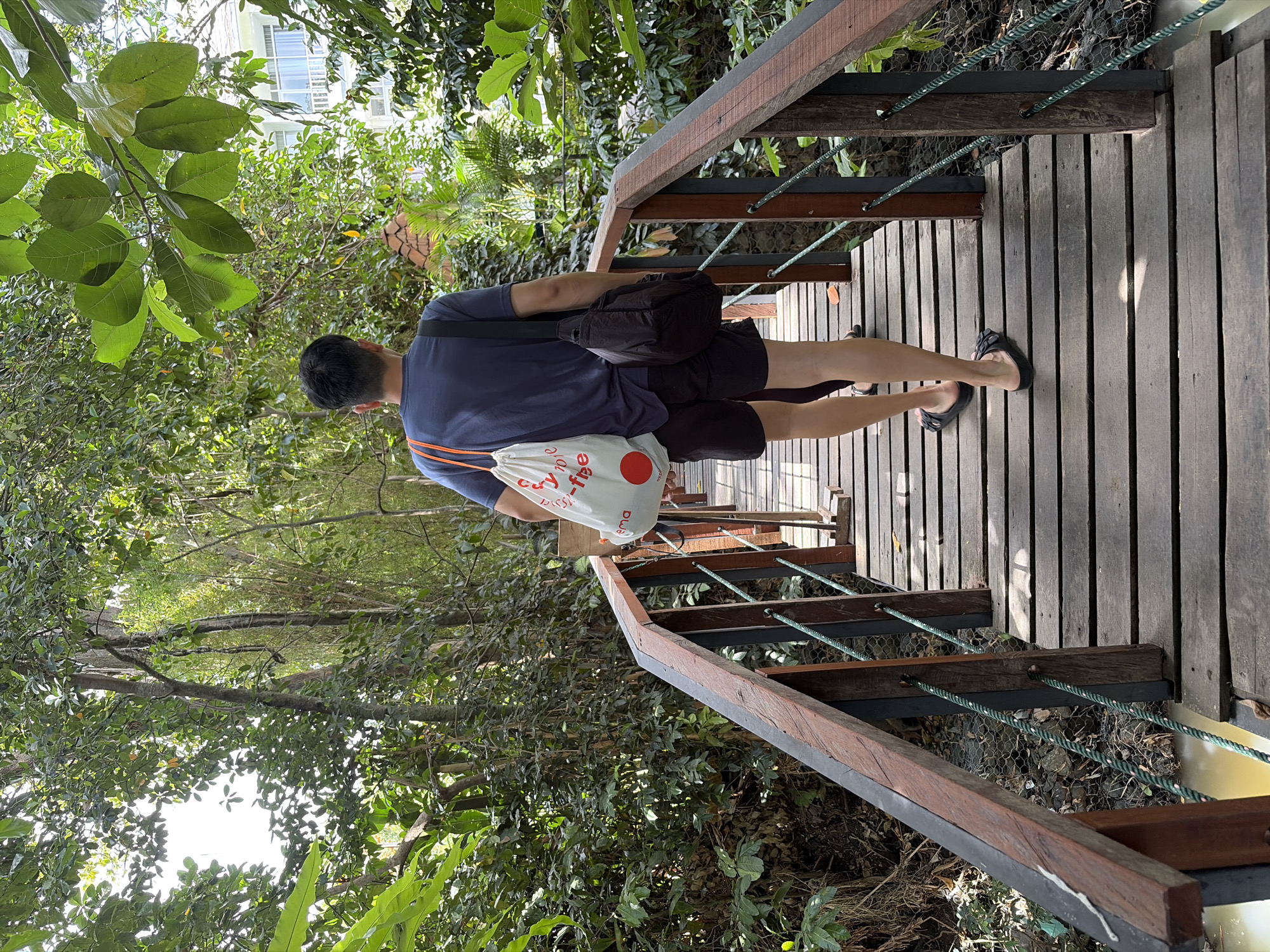 A man walks away from the camera on a wooden bridge in a lush, green environment. He is wearing a dark blue t-shirt, black shorts, and black slides. He carries a white tote bag with orange handles over his left shoulder and a black messenger bag over his right shoulder. The bridge is made of wooden planks and has a wooden railing with green rope. The surrounding area is dense with vegetation, including trees and various plants. In the background, a white building with windows is visible through the foliage. The scene appears to be outdoors in a tropical or subtropical setting, possibly in Mengwi, Indonesia, given the dense greenery and architecture.