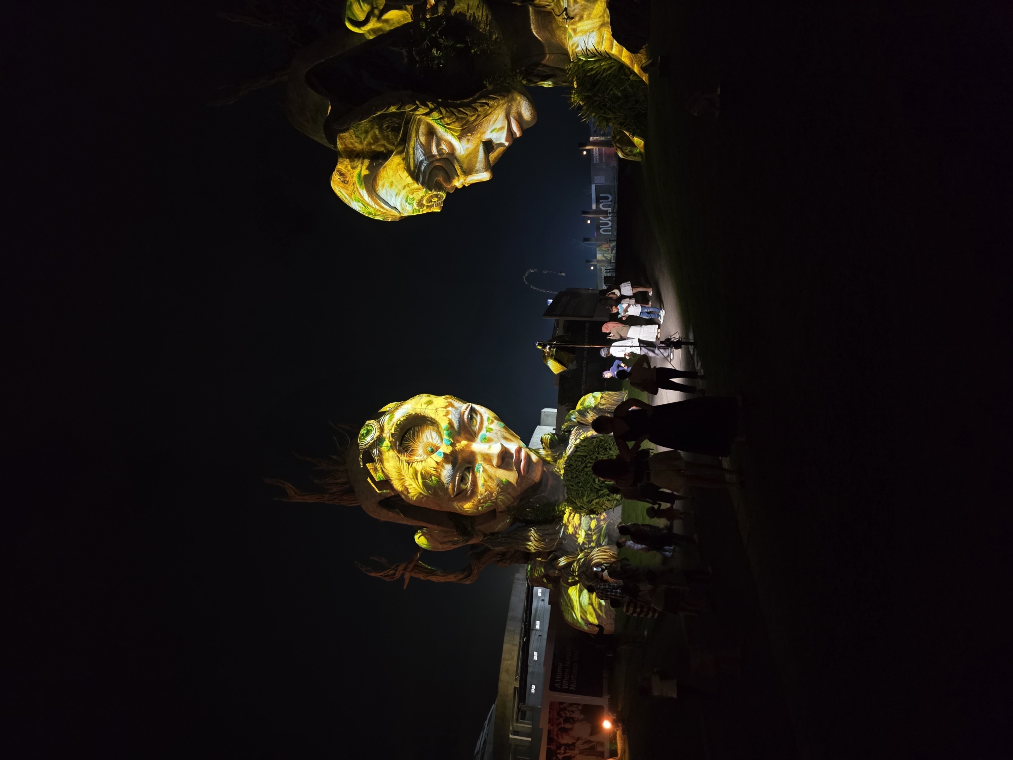At night in Mengwi, Indonesia, an art installation features two large projected faces. A crowd of people stand on a lawn, observing the display. Buildings, walls, and promotional posters can be seen in the background. The faces glow with yellow, green, and blue light.