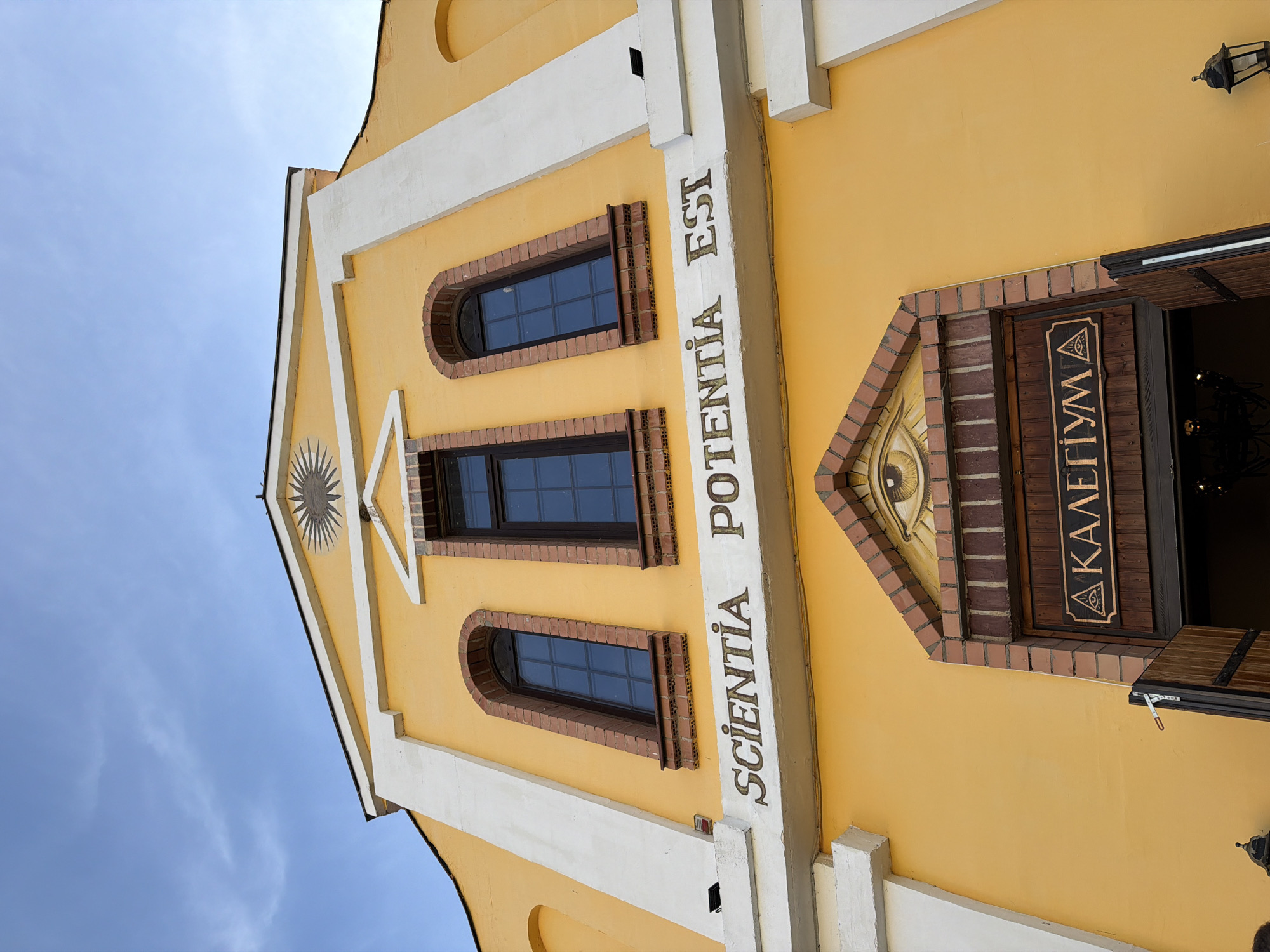 The image shows the facade of a yellow building with a white trim under a bright blue sky with sparse clouds. The building has a triangular pediment at the top with a sunburst-like design. Below the pediment are three arched windows with dark panes, framed by brickwork.
A white band runs across the facade, inscribed with the Latin phrase "SCIENTIA POTENTIA EST" in gold lettering.
Below this, a wooden doorway is partially open, revealing a glimpse of a chandelier inside. Above the doorway, a brick arch frames a carving of an all-seeing eye within a triangle. A wooden sign with a similar eye symbol and text in a Cyrillic-like script, which appears to be "Α ΚΑΛΕΓΙΥΜ", is mounted on the door.
The overall impression is that of an old, possibly historical building, with architectural elements that might suggest a fraternal organization or a place of learning. The location in Літвенскі сельскі Савет, Belarus, is not visually evident in the image itself, but the text on the sign could potentially offer clues to its specific function or affiliation.