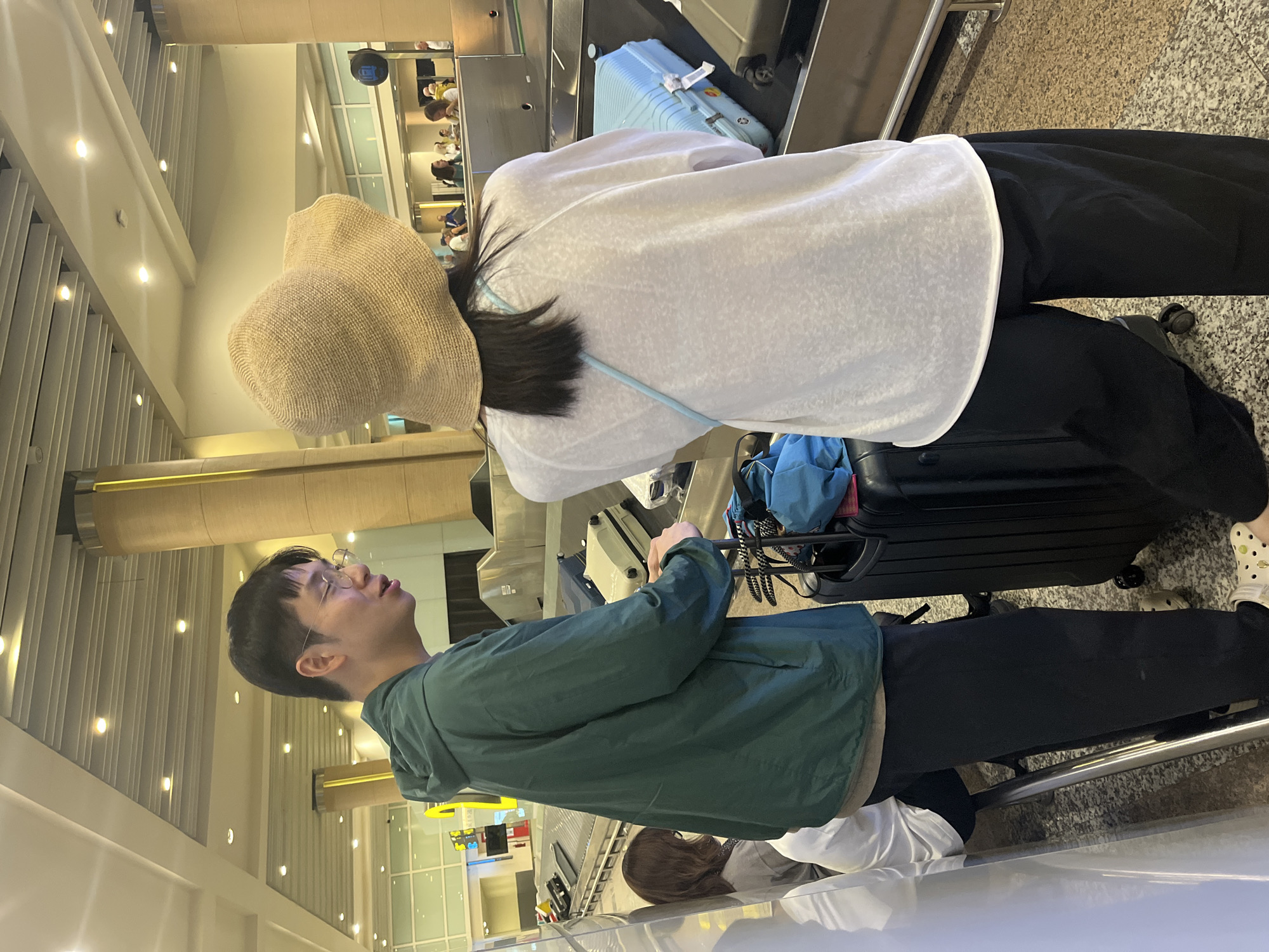 The image depicts an indoor scene at what appears to be an airport baggage handling or check-in area, identified by the request as being in the city of Kuta, Indonesia. In the foreground, two individuals stand beside a baggage conveyor system.
On the left, a young adult male with short dark hair and clear-rimmed eyeglasses wears a dark green hooded jacket over a lighter underlayer, dark pants, and white Crocs-style shoes with dark speckled patterns. His mouth is slightly open, suggesting he is speaking or reacting. His right hand rests on the extended handle of a black hard-shell suitcase at his feet, which is topped with a blue soft-sided bag.
To the right, a second individual, a young adult female, is observed from behind, wearing a large, light brown straw sun hat that covers her head and face. Long, dark hair is visible beneath the hat. She is dressed in a loose-fitting, sheer white long-sleeve top and dark wide-leg pants. A light blue strap of a small bag crosses her back. A light blue hard-shell suitcase rests on the conveyor belt directly in front of her.
The background features multiple sections of baggage conveyor belts, some carrying various pieces of luggage, including a light beige hard-shell suitcase further down. Several indistinct figures of other travelers or airport personnel are visible in the distance. The environment is brightly illuminated by recessed ceiling lights and linear fixtures, featuring large architectural columns with gold-colored accents, a light-colored speckled tiled floor, and large glass partitions in the background.