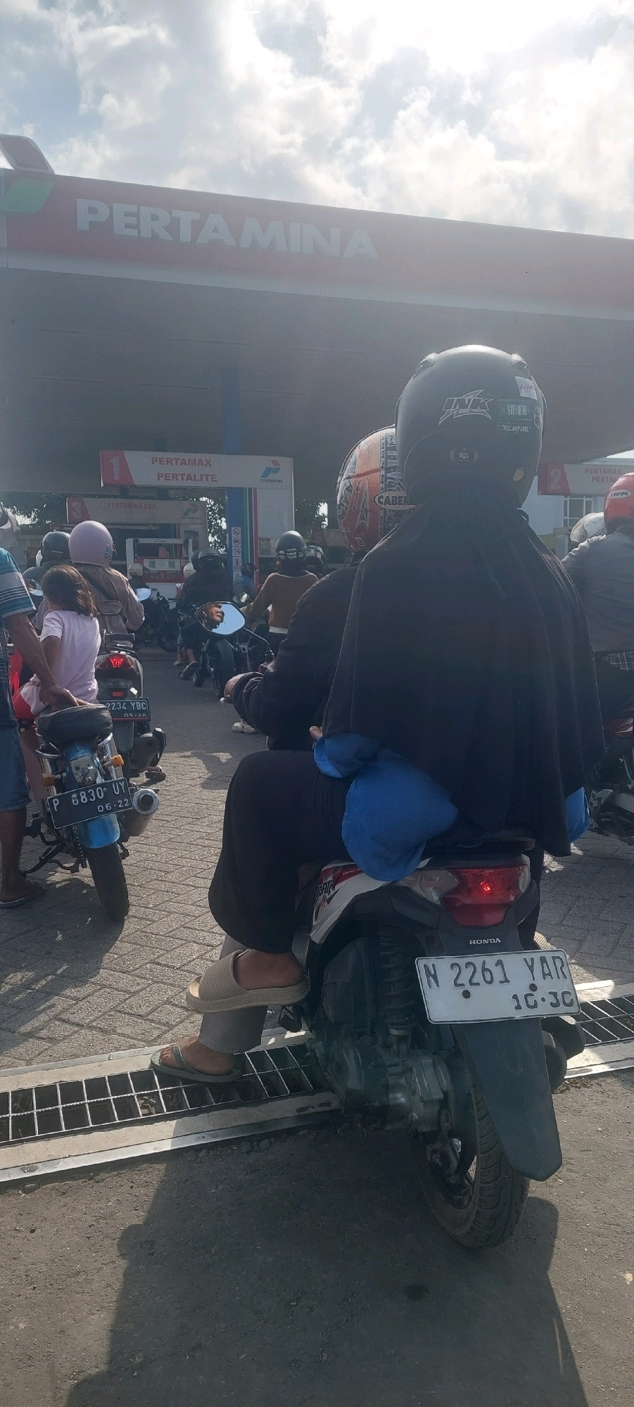 In Lumajang, Indonesia, a queue of motorcycles waits at a Pertamina gas station under a bright, cloudy sky. The foreground shows the back of two motorcycles. The motorcycle on the right has a license plate reading "N 2261 YAR 10-30". A person in dark clothing, wearing a black headscarf and a black jacket over a blue garment, sits on the back of this motorcycle, facing away from the camera. Their feet, clad in light brown slip-on sandals, are visible. The motorcycle on the left has a blue body and a license plate that reads "P 6830 UY 06.22". A person in a pink shirt and a light purple helmet is seated on the back of this motorcycle, facing left. Behind them, another motorcycle with a pink helmet is visible. In the background, the Pertamina gas station canopy is prominent with the word "PERTAMINA" clearly displayed. Signs for "PERTAMAX" and "PERTALITE" are also visible. Other motorcycles and individuals are present, forming a line for refueling. The ground is paved with interlocking bricks in the foreground and asphalt further back.