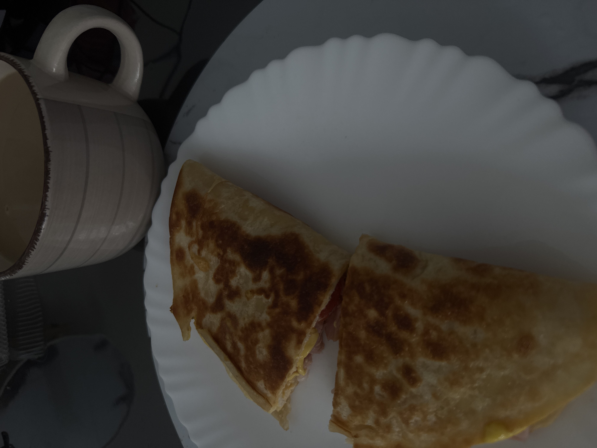 An overhead shot captures a simple meal featuring a quesadilla and a mug, likely set on a table in an indoor setting. The lighting is dim, casting a subdued ambiance over the scene.
In the foreground, a white, round plate with a distinctive wavy or fluted edge holds a quesadilla, which has been cut in half. The quesadilla is golden brown and appears toasted or grilled, indicating it has been cooked. The cut edges reveal its contents, which include melted yellow cheese and what appears to be pinkish meat, possibly ham, or a hint of red vegetable like tomato.
To the left of the plate, a light-colored, possibly ceramic, mug with a prominent handle and vertical striped detailing is visible. The mug contains a light-colored beverage, which could be coffee with milk, tea, or another similar drink. The mug's rim shows a slightly darker, rougher texture.
The background is dark and out of focus, providing no specific details about the surroundings but suggesting a clean, uncluttered surface. There are no people visible in the image, and no visible text can be deciphered. The overall presentation suggests a casual meal or snack. Given the context of Bugry, Russia, this depicts a common indoor dining moment.