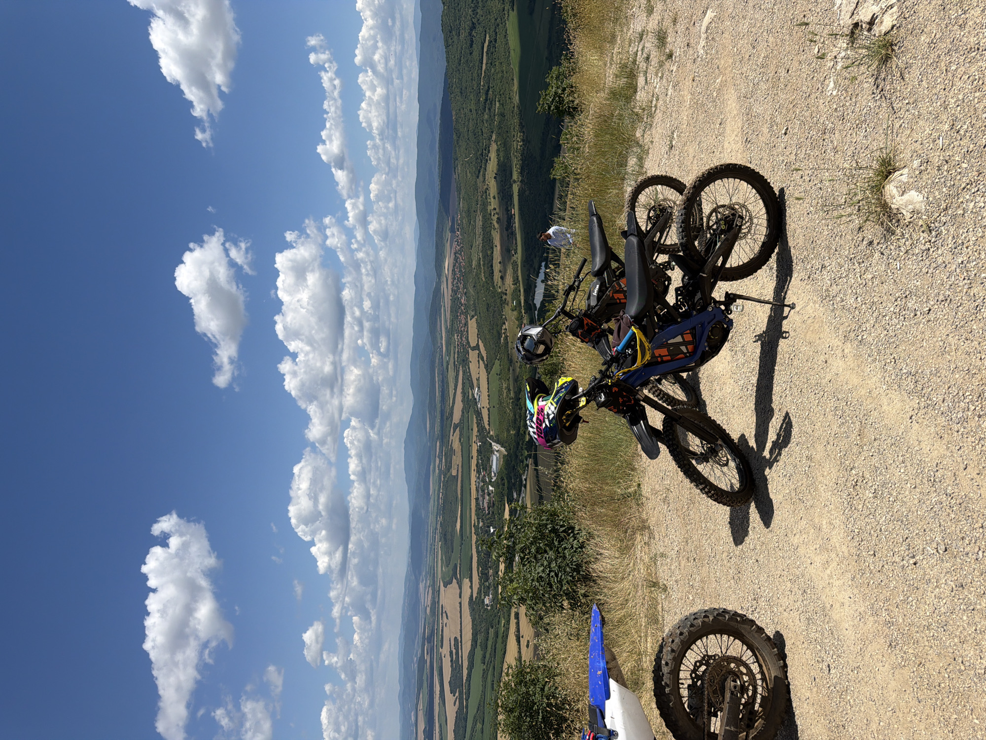 The image captures an outdoor scene with two dirt bikes parked on a gravelly path overlooking a vast, rolling landscape. The sky above is a bright blue, dotted with fluffy white clouds. In the foreground, the rear wheel of a white and blue dirt bike is visible on the left. To the right, a blue dirt bike is parked with two helmets resting on its handlebars, one colorful and the other black. In the background, the terrain stretches out with fields, scattered trees, and a distant town or village. A single person, wearing a white shirt and dark pants, stands in the middle ground, looking out at the scenery. The environment appears to be a hilly, rural area, likely during the daytime with good weather. The location is identified as being in the city of Sheremetia, Bulgaria.