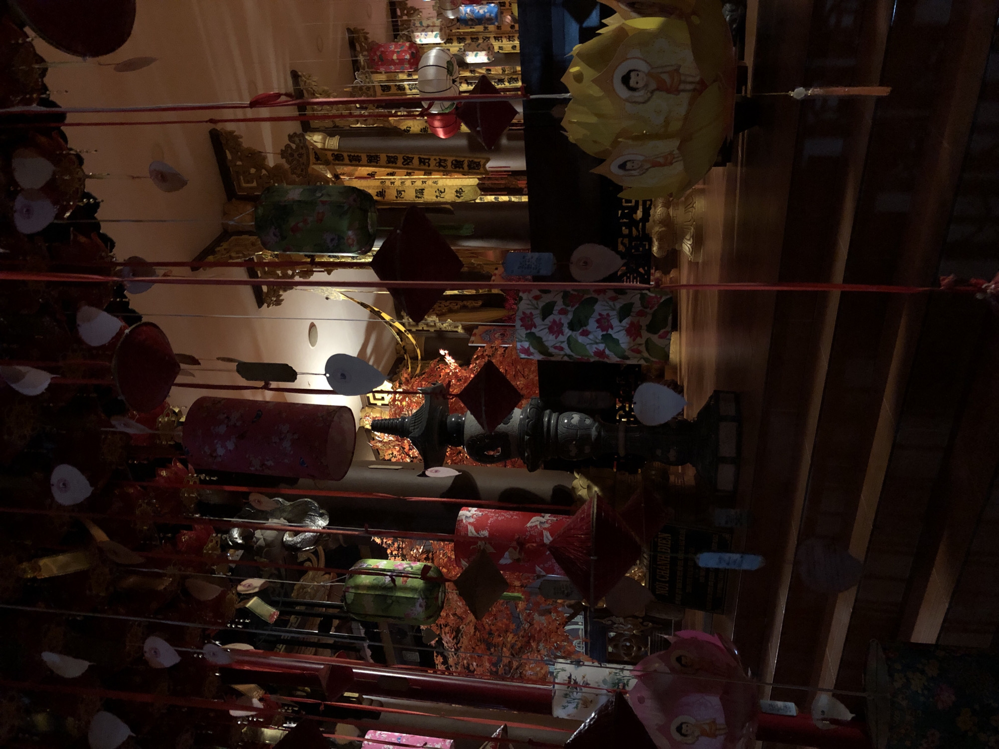 This media file displays a collection of colorful decorative items, possibly for a celebration or market, suspended by red strings. The objects include cylindrical lanterns with floral and crane patterns, square-shaped ornaments, and possibly paper decorations. Some items are illuminated, while others are in shadow, creating a visually dynamic composition.
The scene appears to be indoors, with wooden beams or shelves visible in the background, suggesting a shop or a decorated interior space. The lighting is warm and atmospheric, with some areas brightly lit and others in deeper shadow. There are no people visible in the frame, and the focus is entirely on the decorations.
The overall impression is one of vibrancy and festive decoration. While the exact event is unclear, the abundance and variety of the hanging ornaments suggest a cultural or celebratory context. The setting hints at an Asian location, possibly a market or a shop selling traditional crafts. One piece of text is visible, though blurry, that appears to be in Vietnamese.