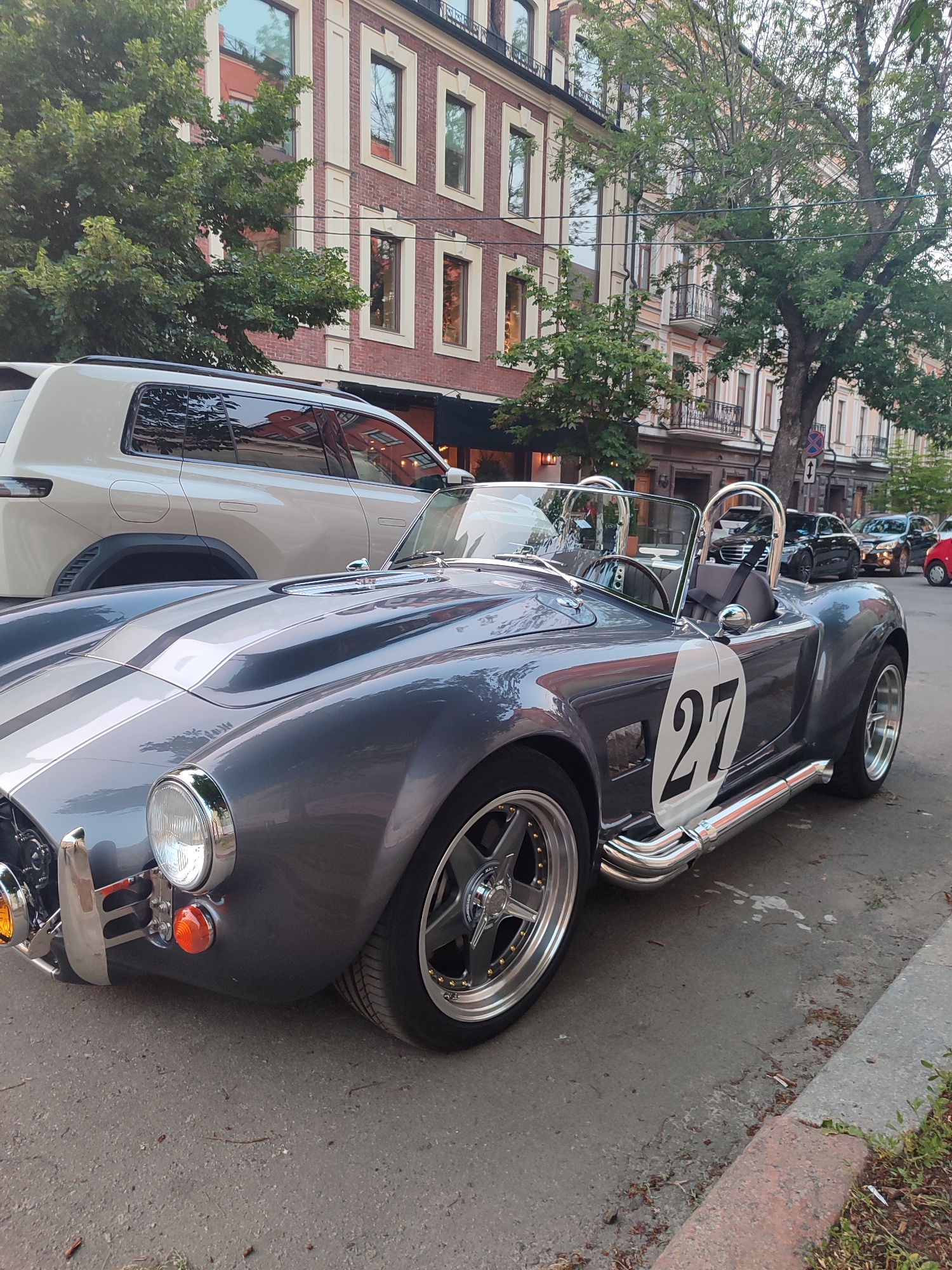 This is a photograph of a grey, open-top sports car with silver racing stripes parked on a street. The car has the number "27" in a white circle on its side. It has a chrome bumper and side exhaust pipes. To the left of the sports car is a white SUV. In the background, there are multi-story buildings with brick and cream-colored facades. Trees with green foliage are visible in front of the buildings. The sky appears overcast, suggesting it might be daytime. The car is the primary subject, and the overall scene depicts a street with vehicles and buildings. There is no visible text other than the number on the car.