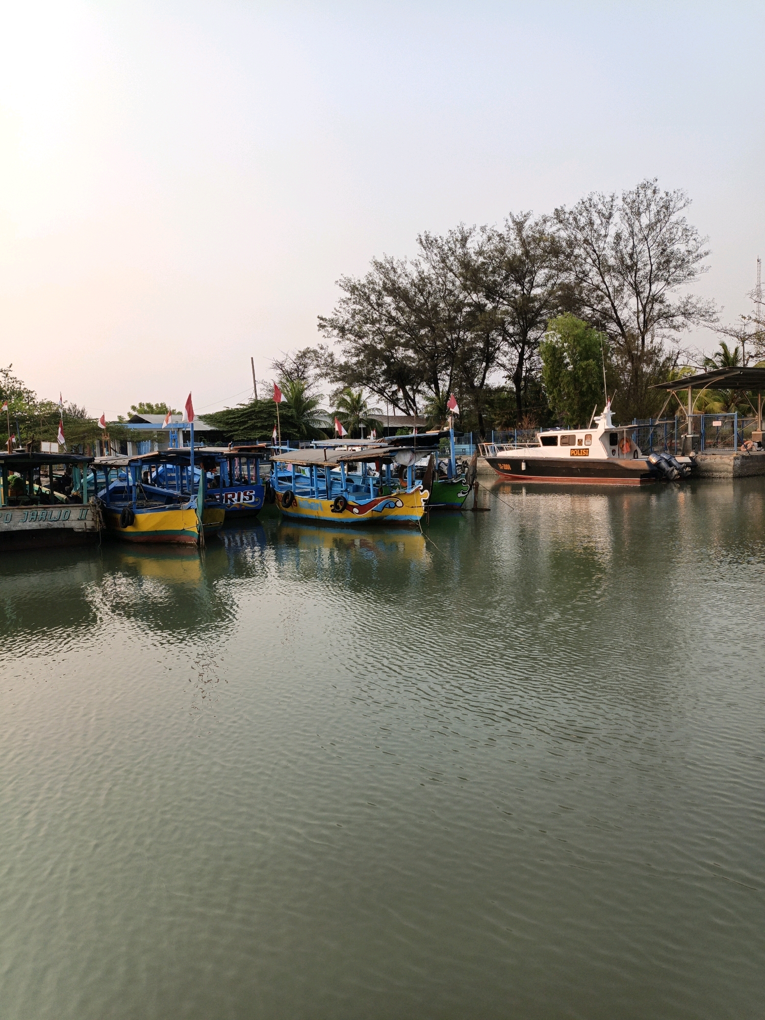 The image shows a waterfront scene in Cikal, Indonesia, likely a harbor or river. Several colorful fishing boats are docked side-by-side in the foreground, their reflections shimmering on the water's surface. The boats are various shades of blue, yellow, and green, with some displaying small red and white flags. Behind the boats, a larger white and black boat with "POLIST" visible on its side is also moored, suggesting a police or patrol vessel. Trees line the background, with some buildings and structures visible behind them. The sky is overcast with a pale, diffused light, indicating either early morning or late afternoon. The water is calm with gentle ripples, and the overall atmosphere is peaceful and serene.