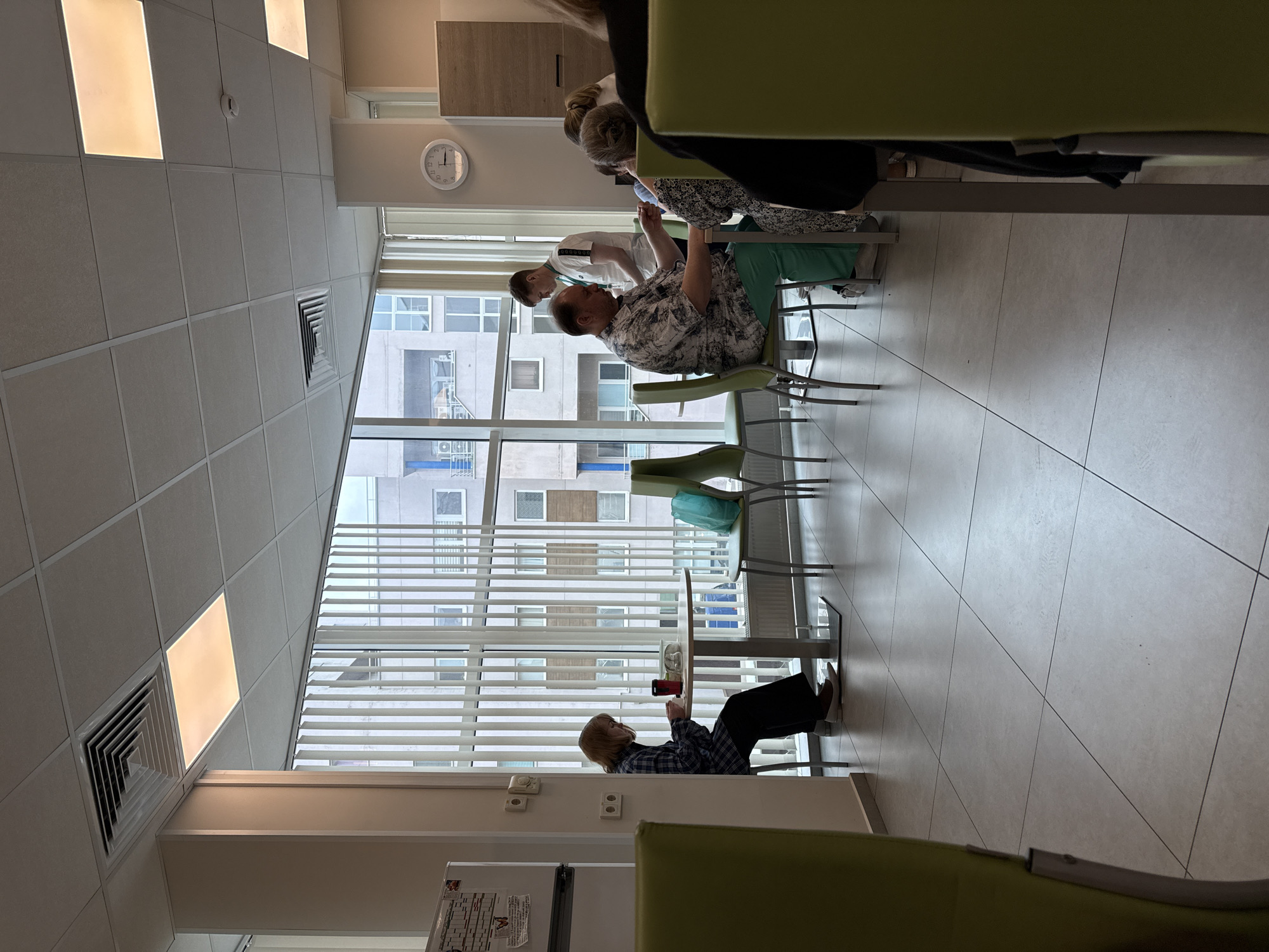 This image captures a bright, modern interior, likely a waiting area or cafeteria in a clinic or public building in Moscow, Russia. The room is characterized by a large, floor-to-ceiling window covering one wall, fitted with horizontal blinds, through which several multi-story buildings are visible.
Several individuals are present. In the mid-ground, a person with blonde hair, wearing a plaid shirt and dark trousers, is seated alone at a small round table, engrossed in their mobile phone. Further back, a man in a patterned short-sleeved shirt is seated at another table, appearing to interact with a person standing beside him. This standing individual wears a white uniform and an ID badge, suggesting they might be staff. Another person with dark hair is partially visible, seated at the same table as the man.
The room features a light-colored tiled floor and a suspended ceiling composed of square white panels, some of which are illuminated as light fixtures. An air vent and a fire alarm are visible on the ceiling. A circular wall clock, positioned high on the wall near the window, shows the time to be approximately 12:45. The furniture includes small round tables and chairs, some with green upholstery and others with metallic frames. On a wall in the lower left, a portion of what appears to be an information board or schedule is visible, but any text on it is illegible. The lighting is bright and diffuse, consistent with daytime. The overall atmosphere appears calm, with people either waiting or resting.