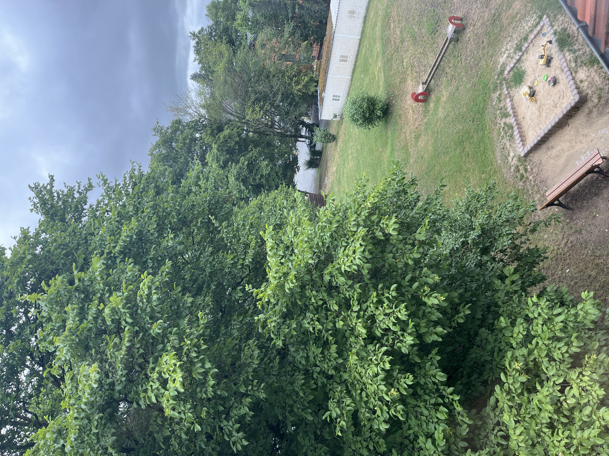 This is an aerial outdoor shot of a small, grassy playground and sandbox area adjacent to a building with a sloped, tiled roof. The playground features a sandbox with several toy construction vehicles, a wooden bench, and a see-saw with red arches. Beyond the playground, there's a grassy lawn with a few small trees and bushes. In the background, a white building with several small windows is visible, along with more trees and buildings, and a road with a parked car. The sky is overcast and cloudy, suggesting a possibly rainy day. The overall scene appears to be located in a residential area, likely a communal space within an apartment complex or a small public park. The image is taken from an elevated perspective, looking down at the playground and surrounding area.