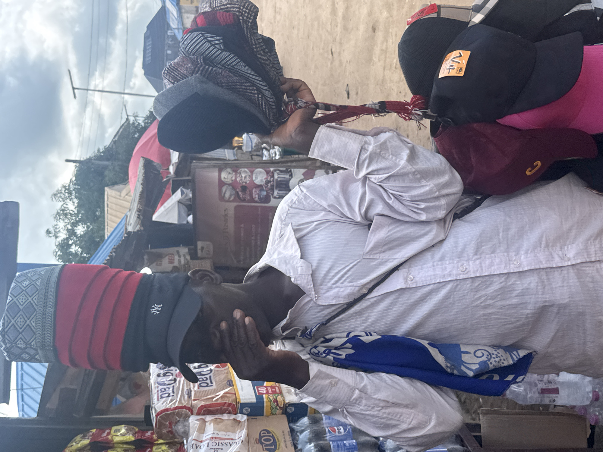 In Ihiagwa, Nigeria, a man stands outdoors at a market stall, his face partially obscured by his hand. He is wearing a long-sleeved, light-colored striped shirt and has a blue and white patterned cloth draped over his shoulder. He balances a tall stack of caps on his head, the topmost cap featuring a gray and black geometric pattern, followed by red and black striped caps, and a black cap with the New York Yankees logo. In his right hand, he holds another collection of caps, including dark-colored ones with patterned fabric and a black baseball cap with an orange tag. Behind him, shelves are stocked with bread loaves and bottles of what appear to be soft drinks. The environment suggests an open-air market with a dirt ground and simple structures with corrugated metal roofs in the background under a cloudy sky.