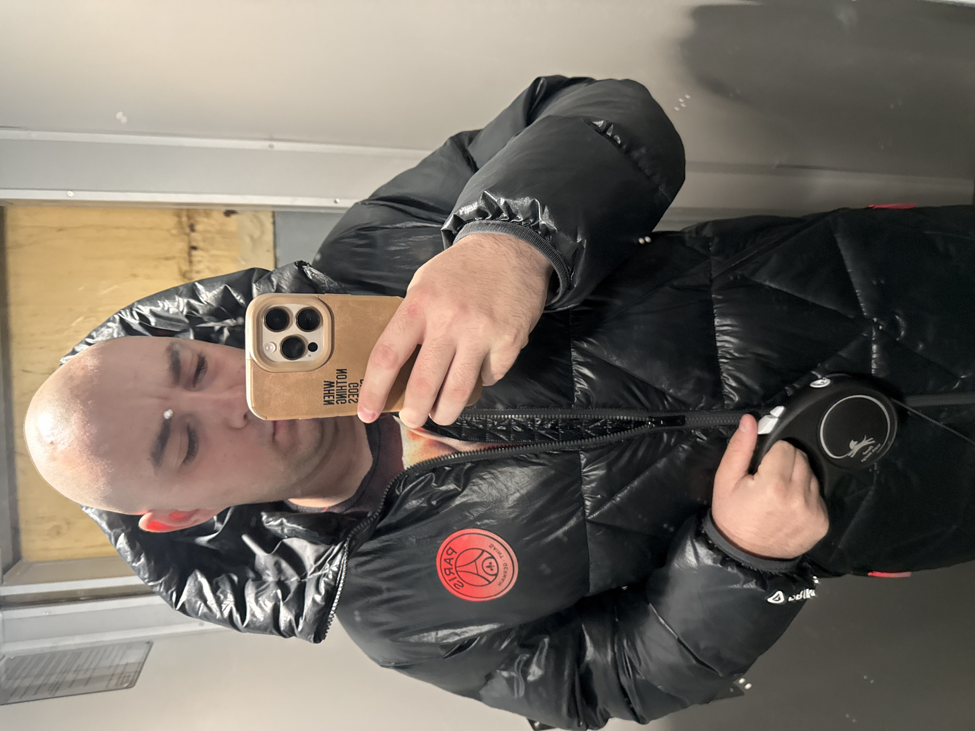 In an interior setting within the city of Pleven, Bulgaria, a bald adult male is depicted in a mirror selfie, his head tilted downwards towards his smartphone. He is wearing a black, shiny, puffer-style winter jacket with a prominent hood, partially unzipped. On the left chest of the jacket, a circular red patch with the "PARIS SAINT-GERMAIN" logo is visible, reversed due to the mirror image. Beneath the unzipped jacket, a patterned shirt is partially visible. The man holds a light tan-colored smartphone with a multi-lens camera system in his right hand, reflecting text on its case that appears as "WHEN NOTHING GOES ZJOK" (reversed for "WHEN NOTHING GOES RIGHT"). In his left hand, he holds a black retractable dog leash, which features a white cat silhouette logo and partially visible text "flexi New Classic Small". The background of the reflection shows a plain, light-colored wall section on the right, and a textured, possibly paneled or wooden surface on the left with vertical lines and a warmer tone. Vertical metallic strips or door frames are also visible in the reflected background. A small white mark is present on the man's forehead.