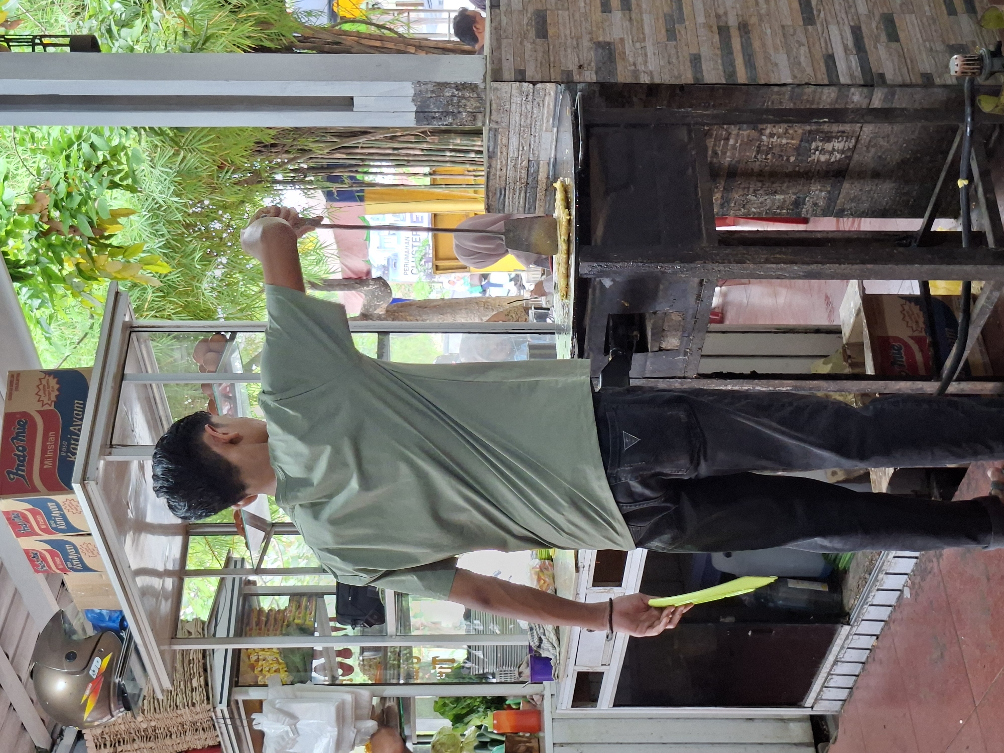 A man in a light green t-shirt and dark pants is operating a food stall. He is standing next to a metal cart with glass displays containing various food items. Behind him is a cooking station with a griddle. Boxes of "Indo Mie" instant noodles are stacked on top of the cart. The scene appears to be outdoors, possibly in Lhokseumawe, Indonesia.