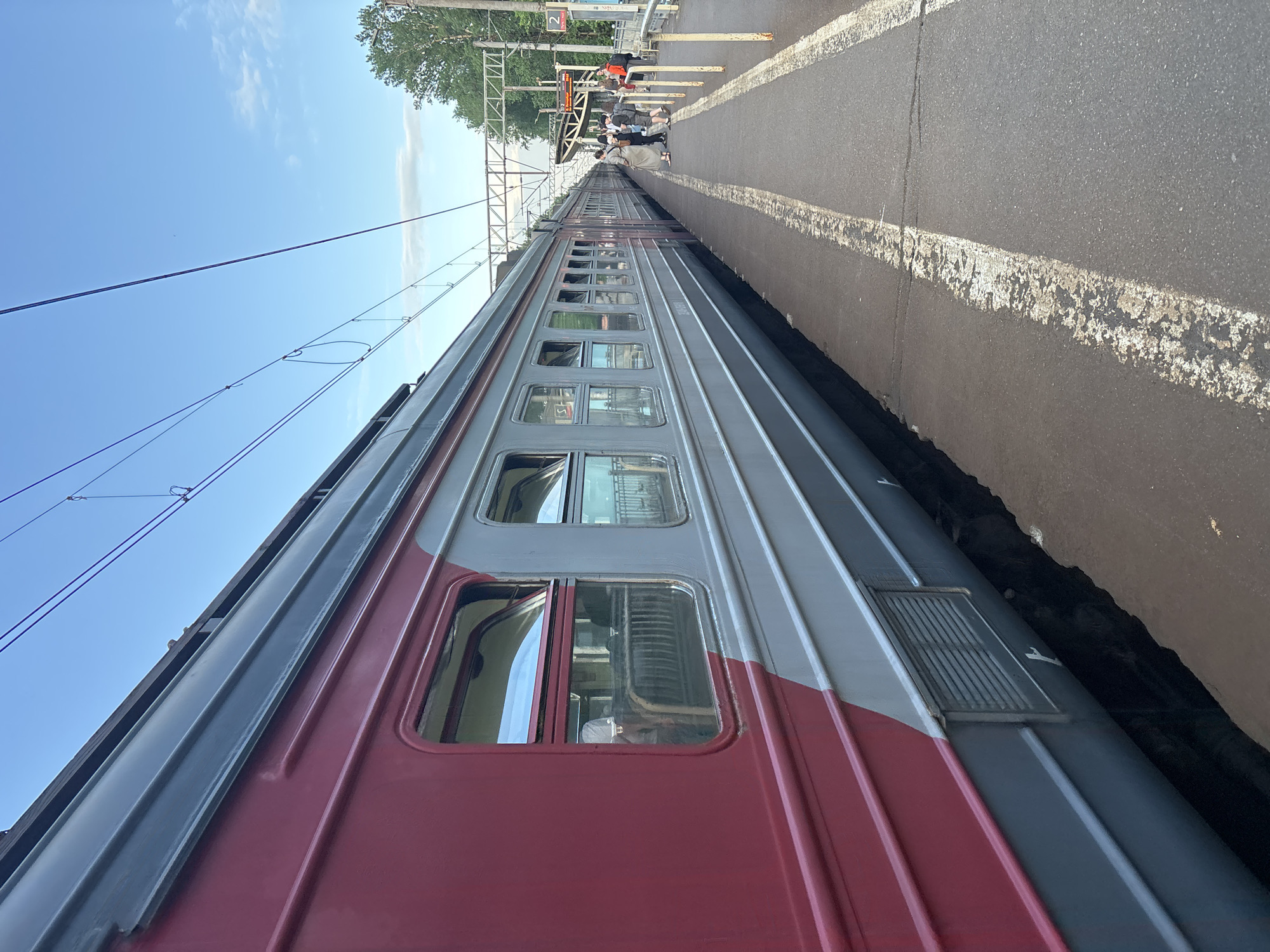 A train is stopped at a platform under a clear blue sky, likely during the daytime. The image, originally rotated, shows the side of a long train consisting of multiple carriages, painted in a two-tone scheme of dark red on the lower half and a lighter grey/silver on the upper half. Numerous windows are visible along the side of the train, some appearing to be open or tilted. A vent is visible on the side of one of the grey/silver carriages near the bottom of the frame.
To the right of the train is an asphalt platform with worn white lines marking its edge. Further down the platform, a group of people is visible, some standing and some possibly seated under a structure that appears to be a shelter or part of a station building. Overhead power lines and their support structures stretch across the sky above the train. Trees with green foliage are visible in the background on the far side of the platform.
A sign with red and white coloring, possibly indicating track number "2", can be seen on a pole above the platform where people are gathered. The setting suggests an active train station in Komarovo, Russia.