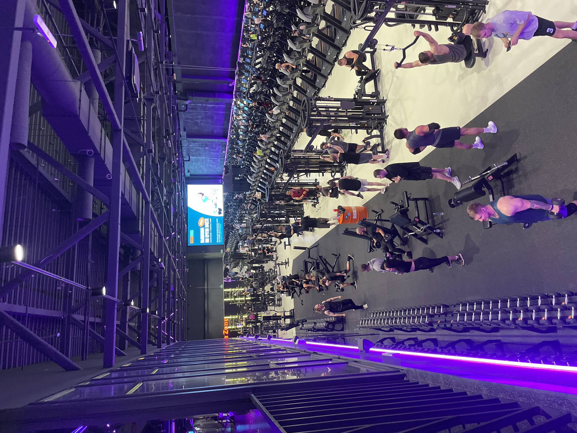 The image captures a well-lit, modern gym interior, dominated by sleek black gym equipment and vibrant purple accent lighting. The scene is bustling with activity, featuring numerous individuals engaged in various workouts. In the foreground, several men are seen lifting weights, some with dumbbells and others on weight machines. The perspective is from a slightly elevated angle, looking across the gym floor.
Further back, rows of treadmills are occupied by people exercising. The gym appears to be spacious, with a high ceiling and industrial-style metal beams and structures. On the left side of the image, a large screen displays an advertisement or informational graphic with text that is partially visible. The overall atmosphere suggests a dynamic and active environment. The lighting is primarily artificial, with purple lights creating a distinct mood, and overhead lights illuminating the workout areas. There are no clear indications of the time of day or weather conditions. The focus is on the physical activities and the gym's modern infrastructure.