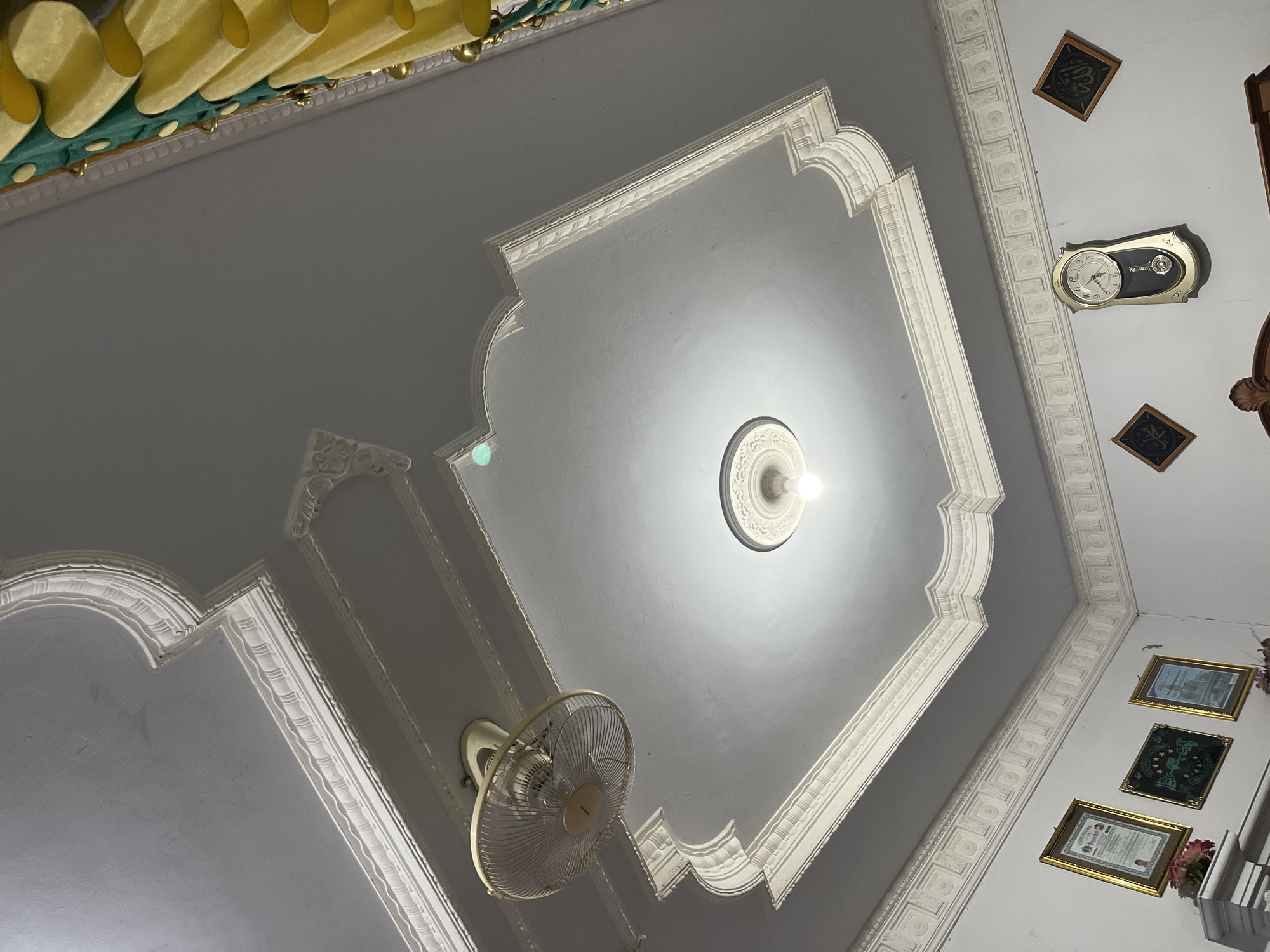 The image captures an interior view of a room, focusing on the ceiling and upper portion of the walls. The ceiling features ornate white plaster molding with decorative details and a central recessed area with a light fixture. A ceiling fan is visible on the left side. The walls are painted white and display several framed items, including certificates and pictures. A pendulum clock is mounted on the right wall, accompanied by two small framed religious inscriptions. Yellow and green drapery with gold accents is visible along the top edge of the image on the right. The overall scene suggests a well-maintained domestic interior. The location is identified as Glagah Kulon, Indonesia.