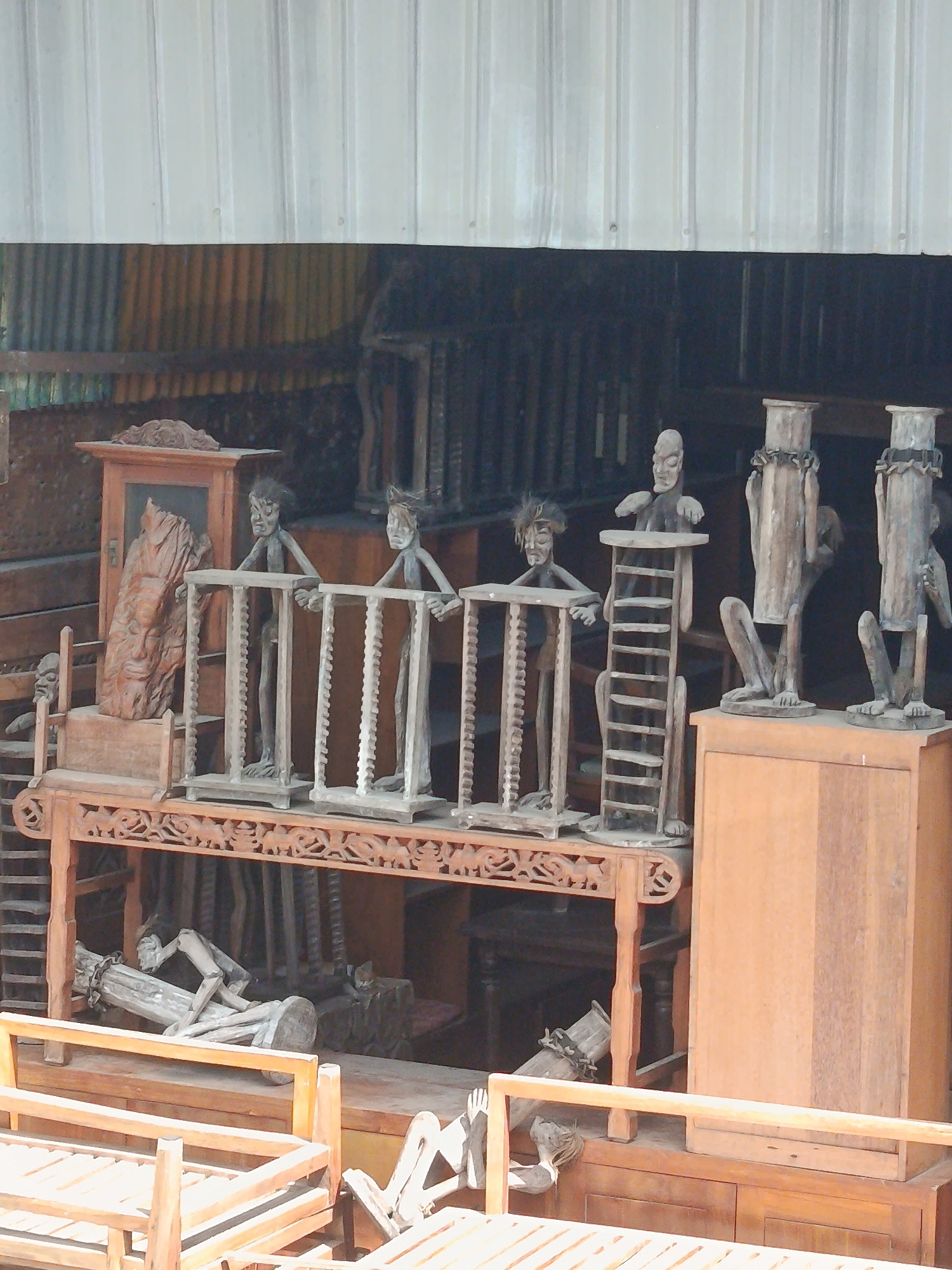 In Kuta, Indonesia, a workshop displays numerous wooden sculptures. The foreground features an arrangement of what appear to be wooden beds or benches, made of light-colored wood with slatted surfaces and simple railings. Behind these, a more intricate wooden structure, possibly a display shelf or platform, holds several carved wooden figures. These figures are stylized, resembling human forms with exaggerated features. Some are thin and elongated, standing behind wooden cages or frames, while others are more abstract, with cylindrical torsos and detached limbs. One prominent sculpture on a separate wooden cabinet is a large, carved wooden relief of a face. The background consists of more wooden furniture and structures, all seemingly in various stages of completion or display, within a covered, open-air space. The overall impression is of a craftsman's studio or an outdoor market stall specializing in traditional wooden art.